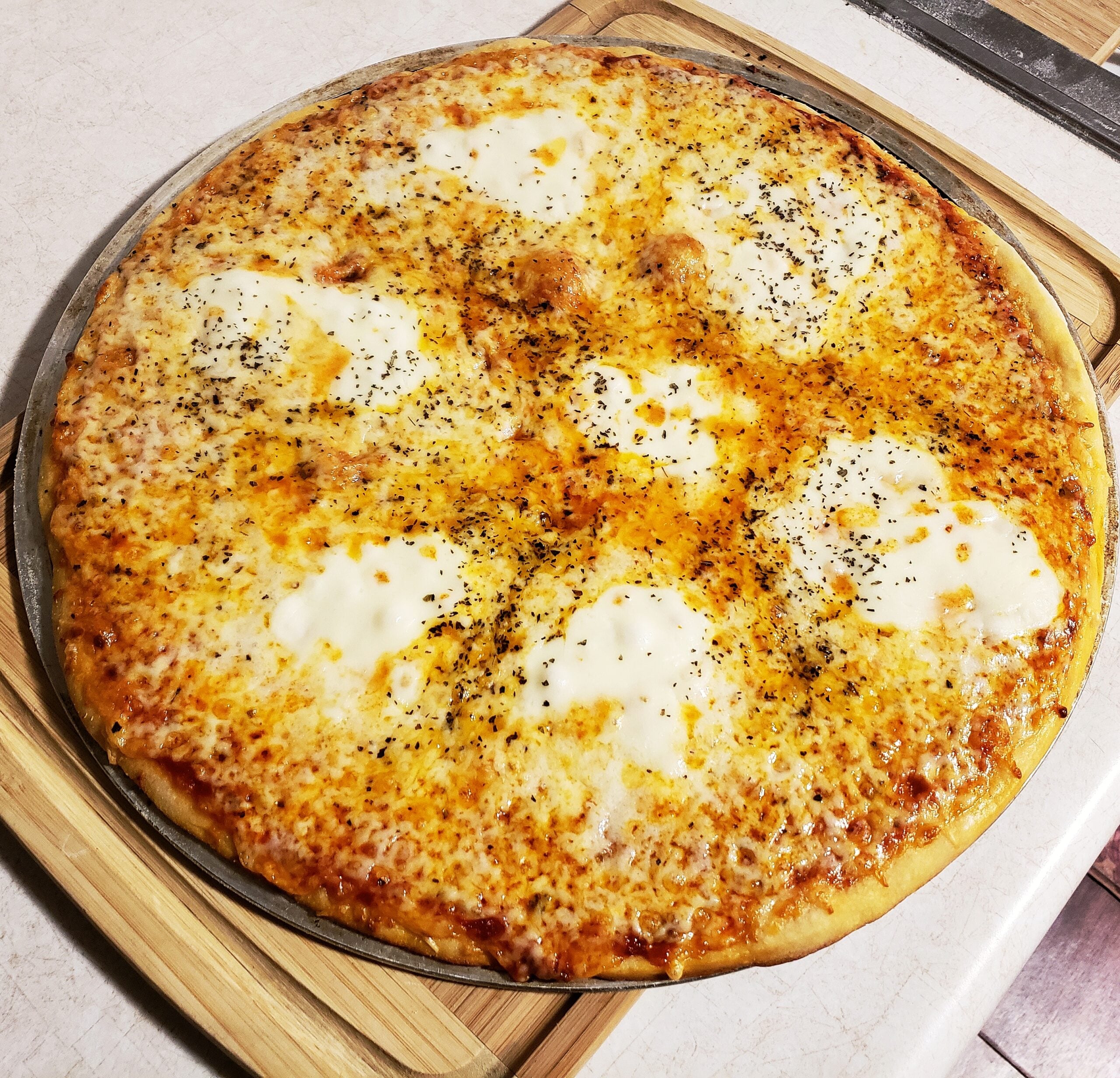 A large cheese pizza with melted mozzarella, tomato sauce, and herbs, displayed on a round metal tray atop a wooden cutting board on a kitchen counter.