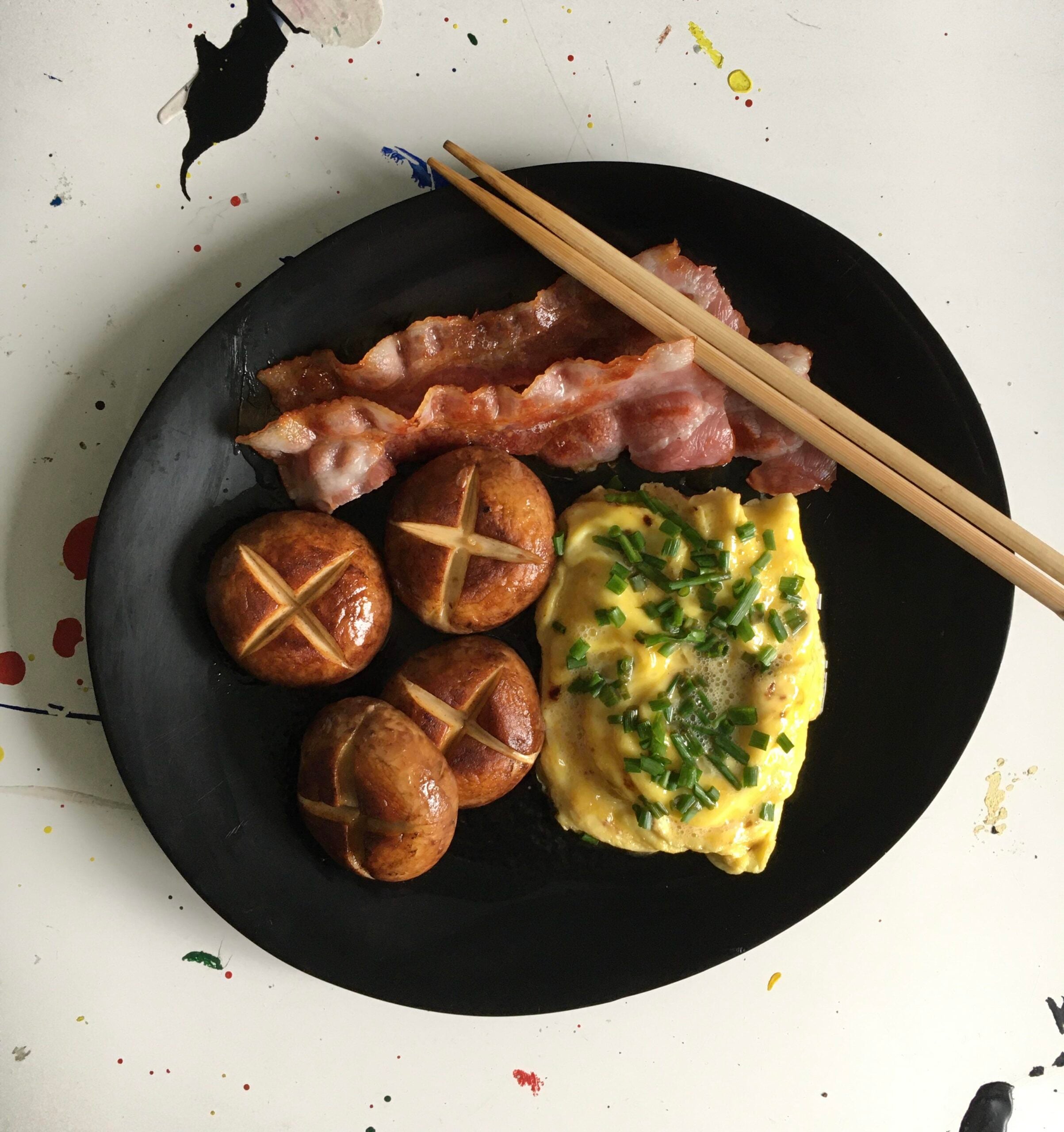 A black plate with scrambled eggs topped with chives, three brown mushrooms, and strips of bacon. A pair of wooden chopsticks rests on the plate. The table underneath has colorful paint splatters.