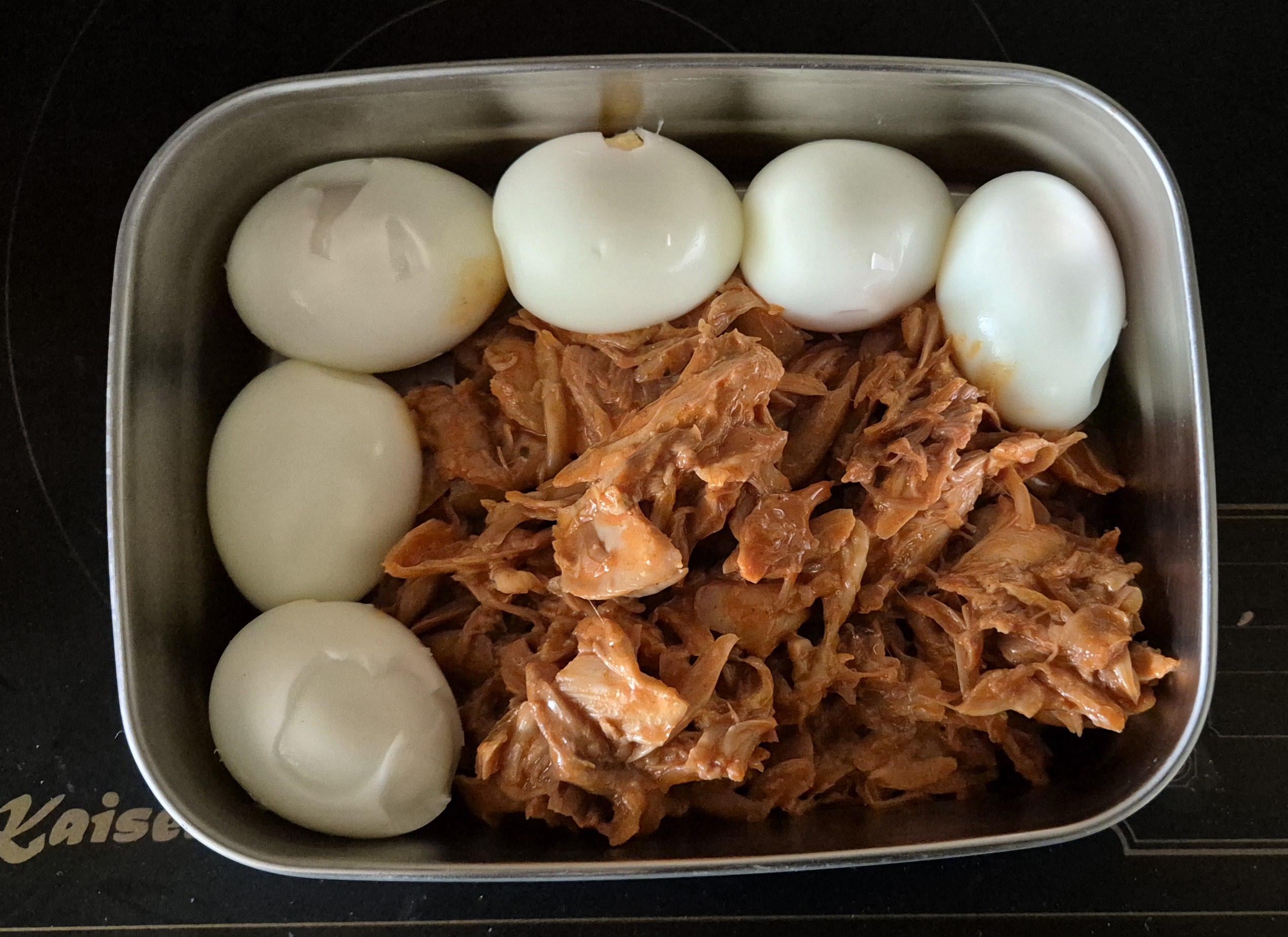 A metal container with six peeled hard-boiled eggs arranged around shredded cooked meat, likely chicken or tuna, sits on a dark surface—an unexpected arrangement that could easily remind one of those hilarious cooking disasters seen online.