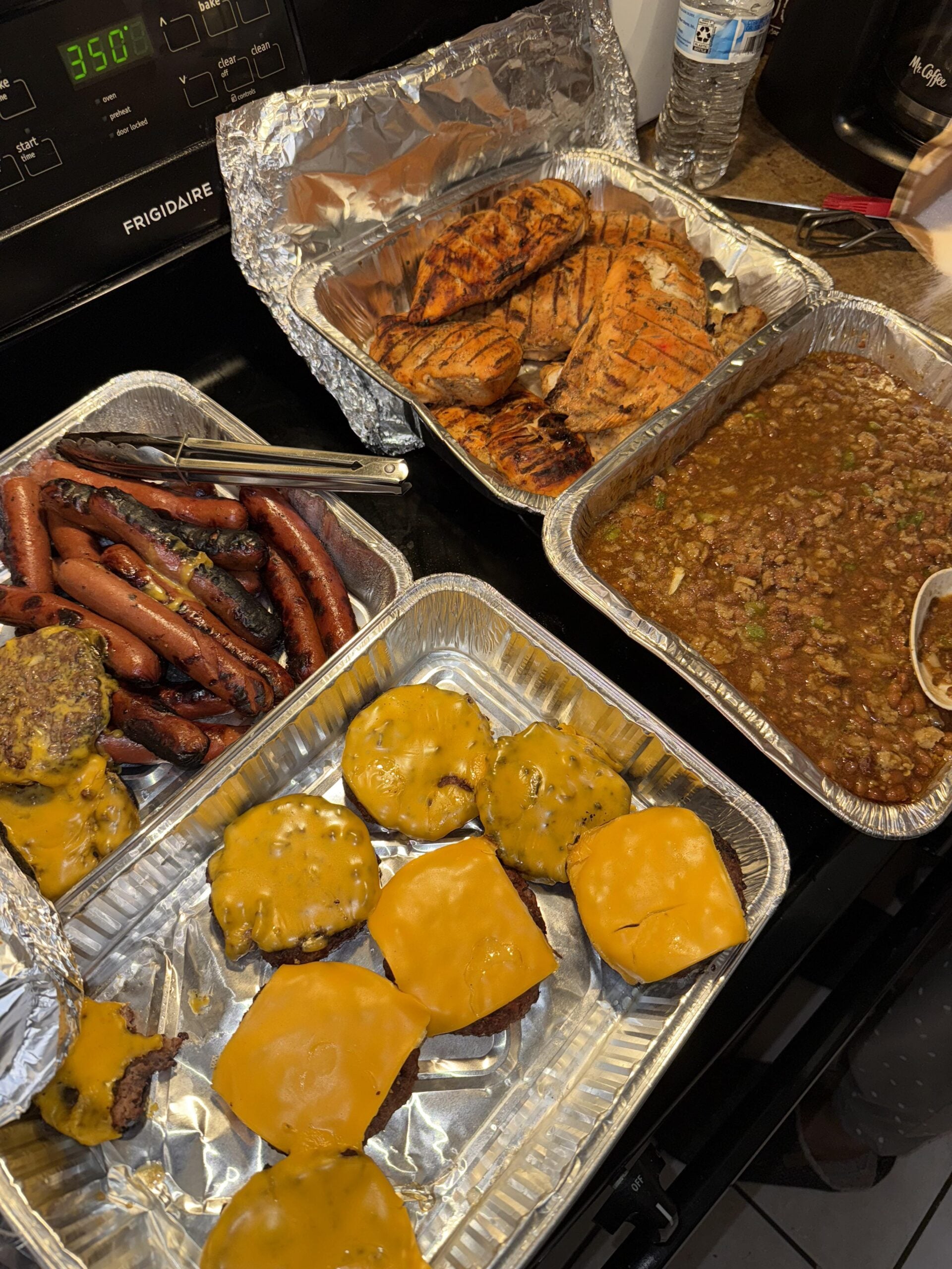 Four trays of food—grilled hot dogs, cheeseburgers, grilled chicken breasts, and baked beans—sit in foil pans on a stove, ready to serve. A water bottle and kitchen utensils are nearby, while the oven display shows 350 degrees. Perfect for the best BBQ cookouts this summer.