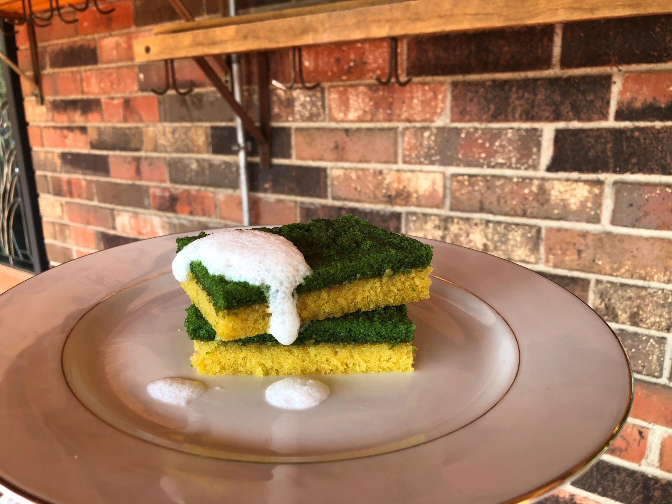 A plated dessert resembling kitchen sponges, with two green-and-yellow layers stacked and topped with white foam, sits on a white plate against a brick wall background.