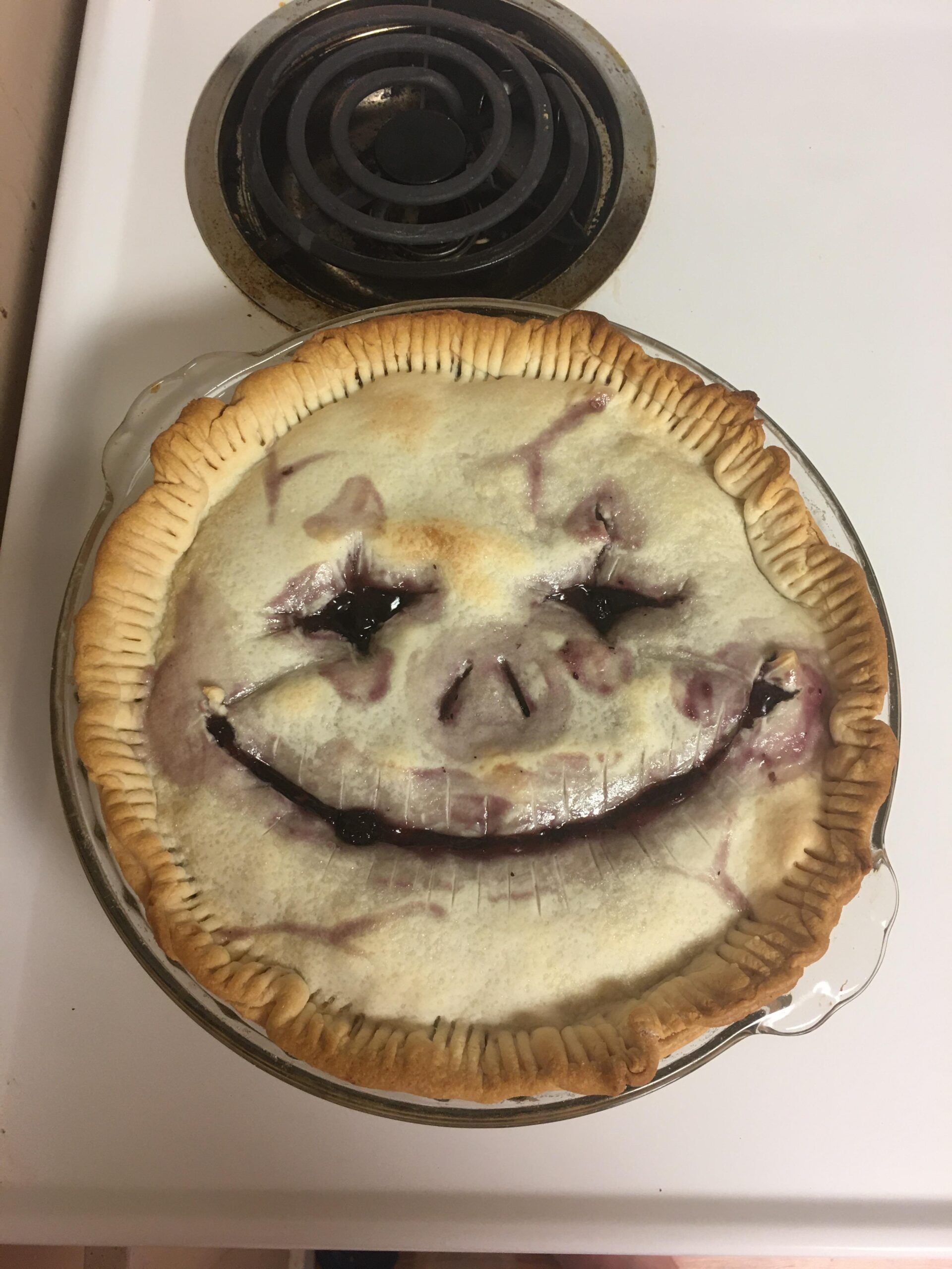 A baked pie with a golden crust has a smiling face drawn with filling, featuring two eyes, a nose, and a wide mouth. The pie sits on a white stovetop near a black coil burner.