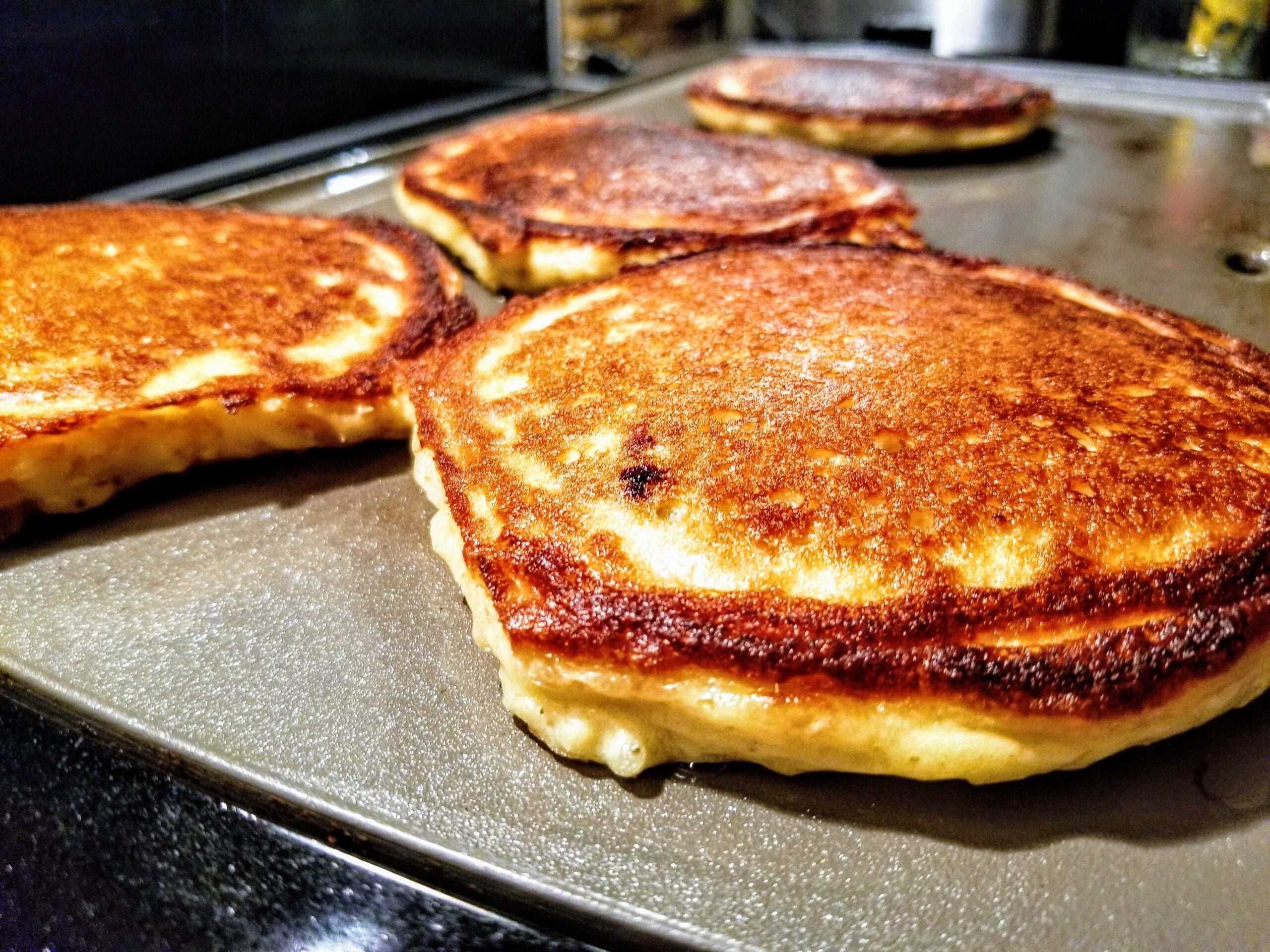 Four homemade pancakes cook on a griddle, their edges crispy and centers fluffy, with a close-up focus on the nearest golden-brown pancake.