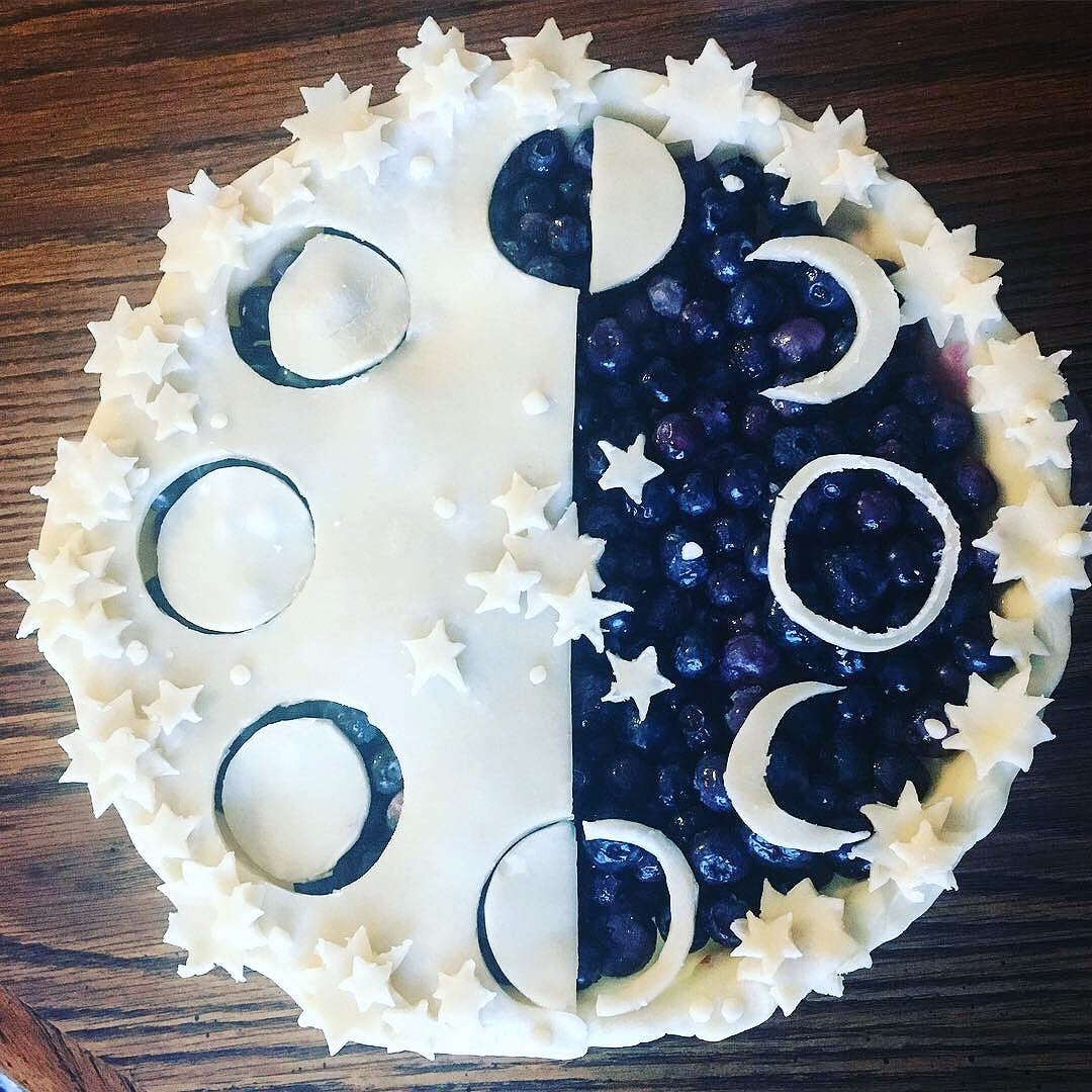 A round pie decorated with pastry cutouts of moons and stars. One half shows exposed blueberries, the other half is covered with white crust, displaying moon phases. The pie sits on a wooden surface.