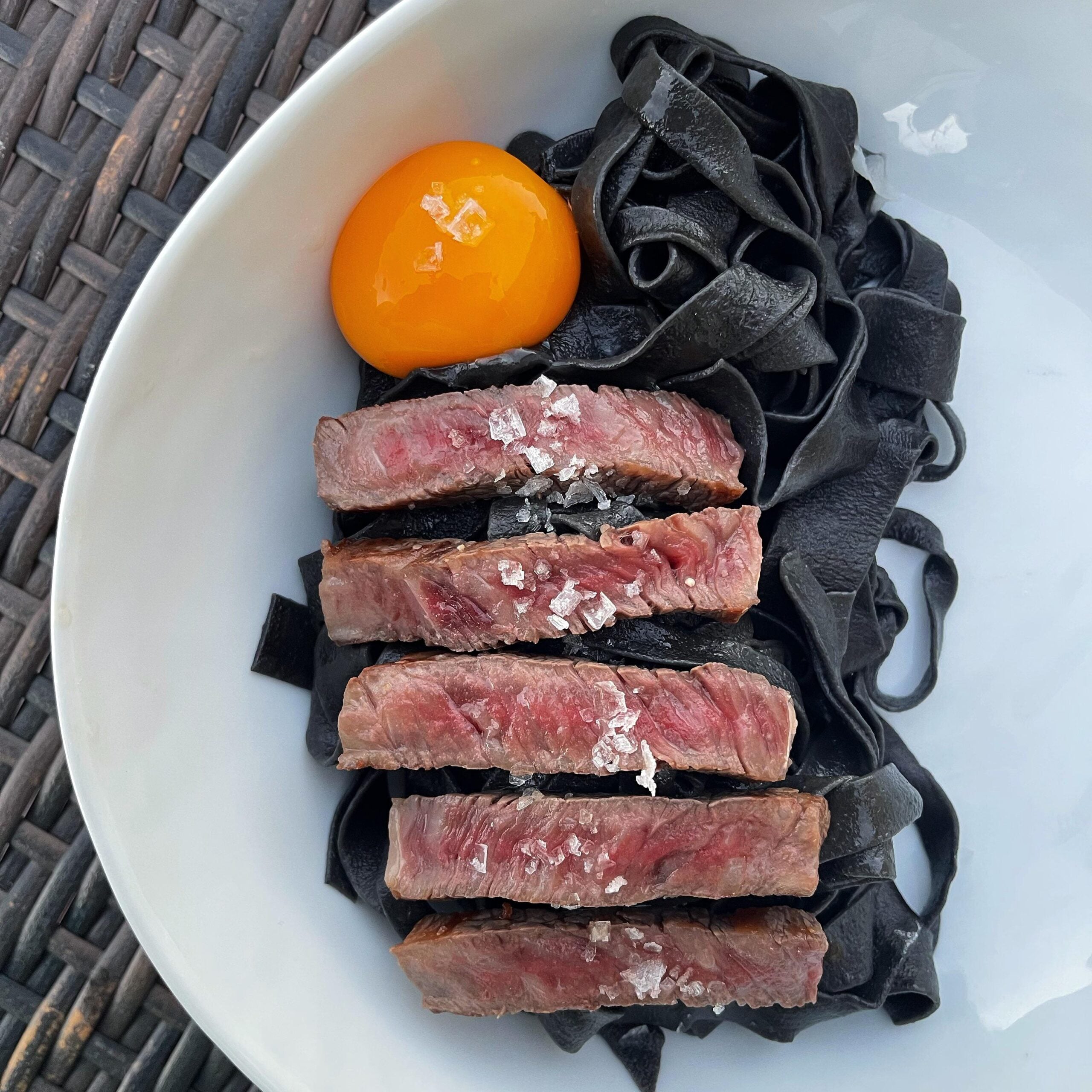 A white bowl with black pasta, five slices of rare steak topped with sea salt, and a raw egg yolk, set on a woven surface.