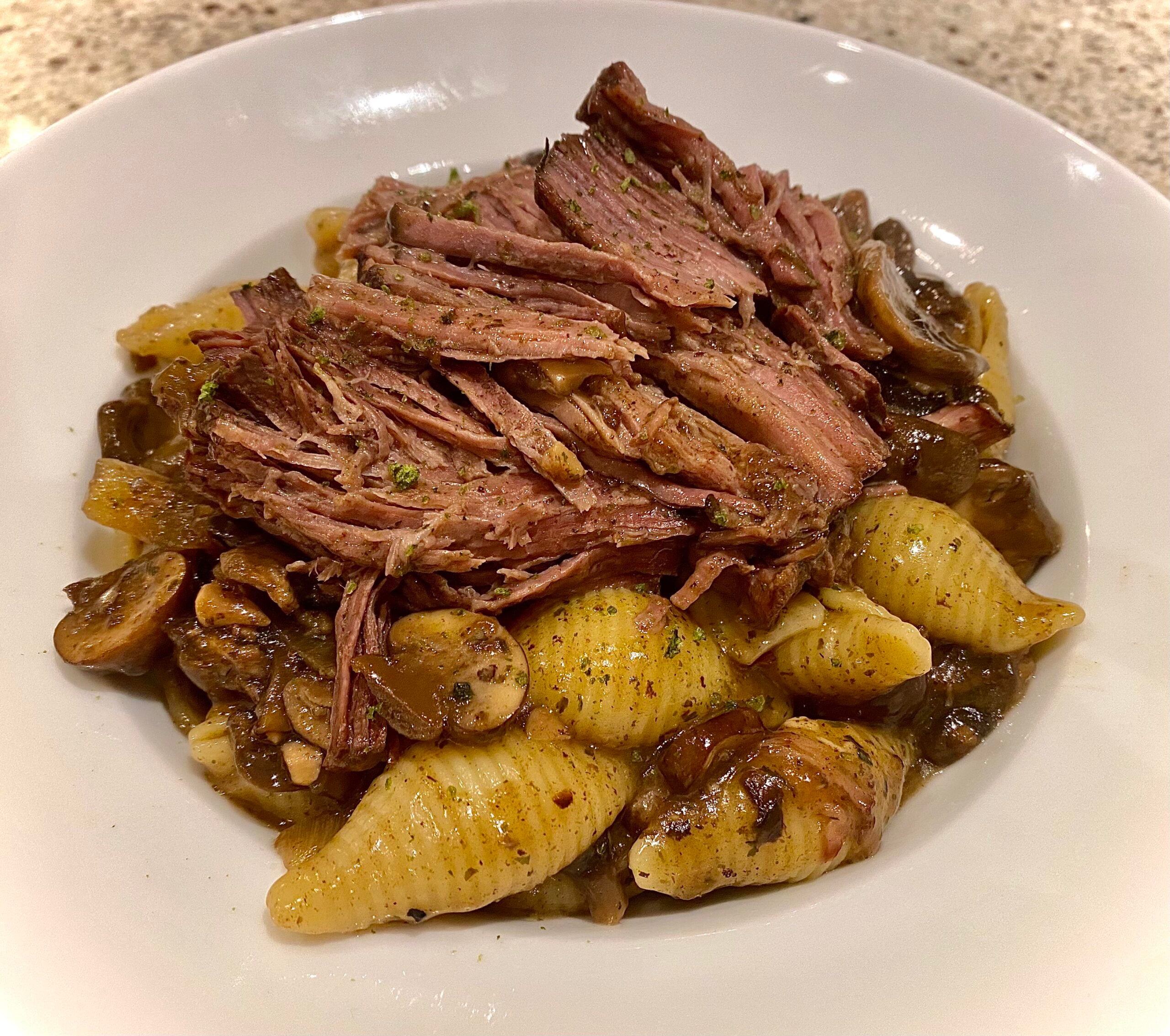 A white bowl filled with pasta shells, sliced mushrooms, and topped with shredded beef, all in a savory sauce.