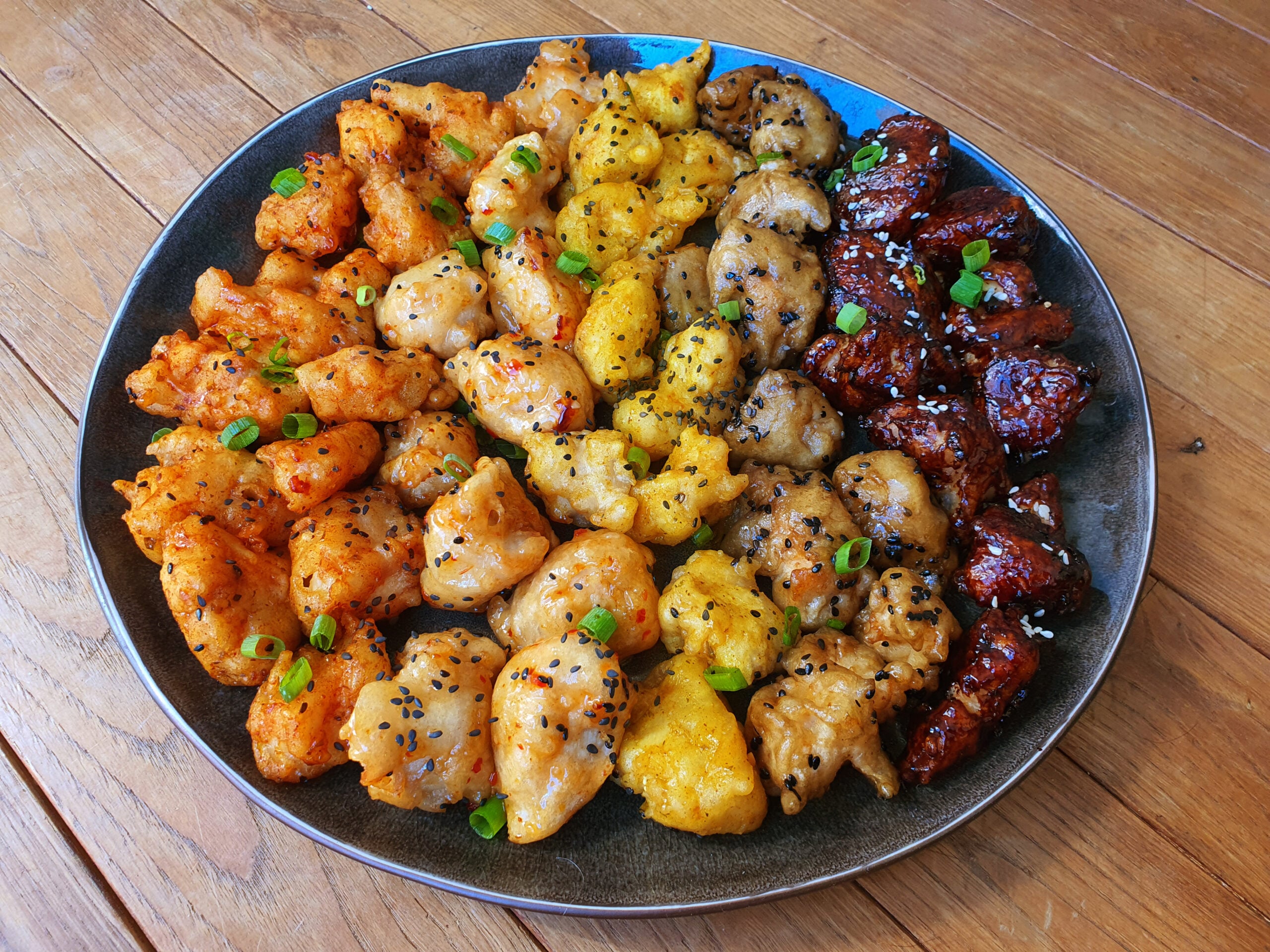 A round plate with colorful bite-sized pieces of fried chicken in various sauces, garnished with green onions and black sesame seeds, arranged neatly on a wooden table.