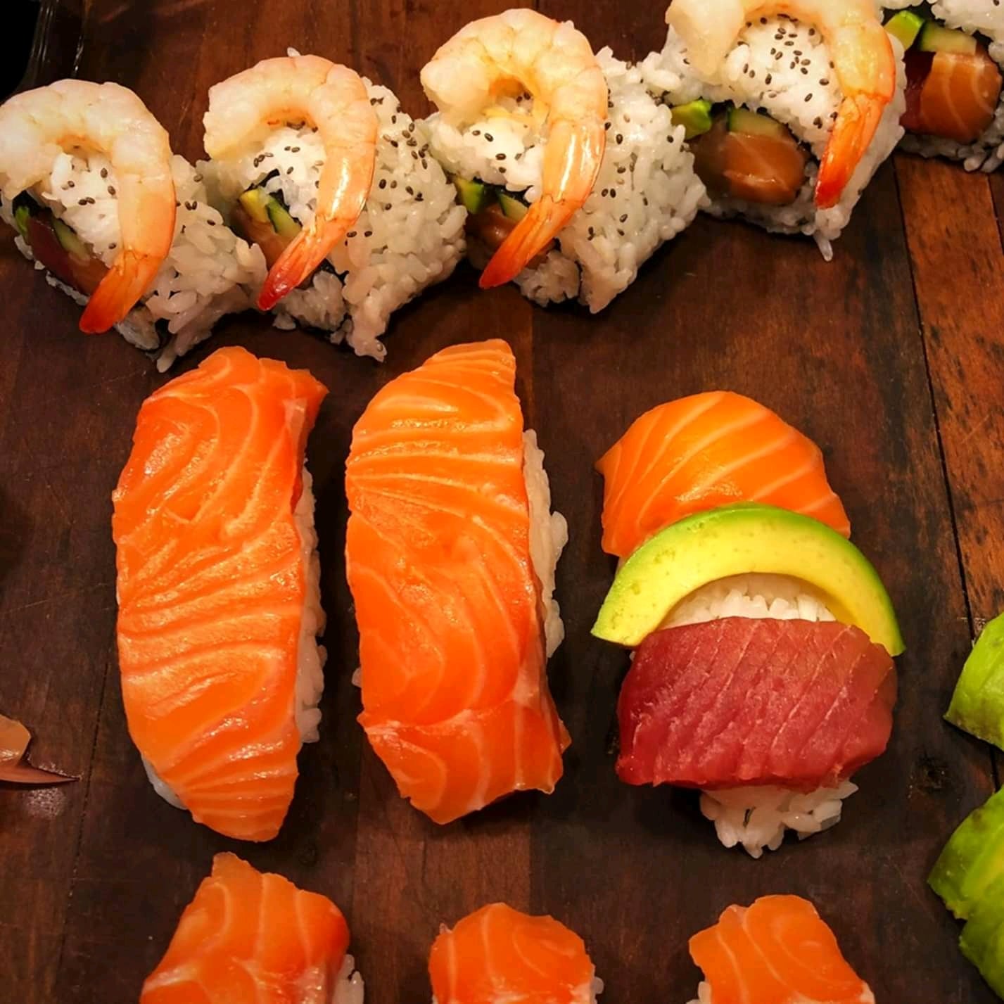 Assorted sushi on a wooden board: shrimp rolls in the back, three pieces of salmon nigiri, and one piece topped with tuna, avocado, and rice in the front.