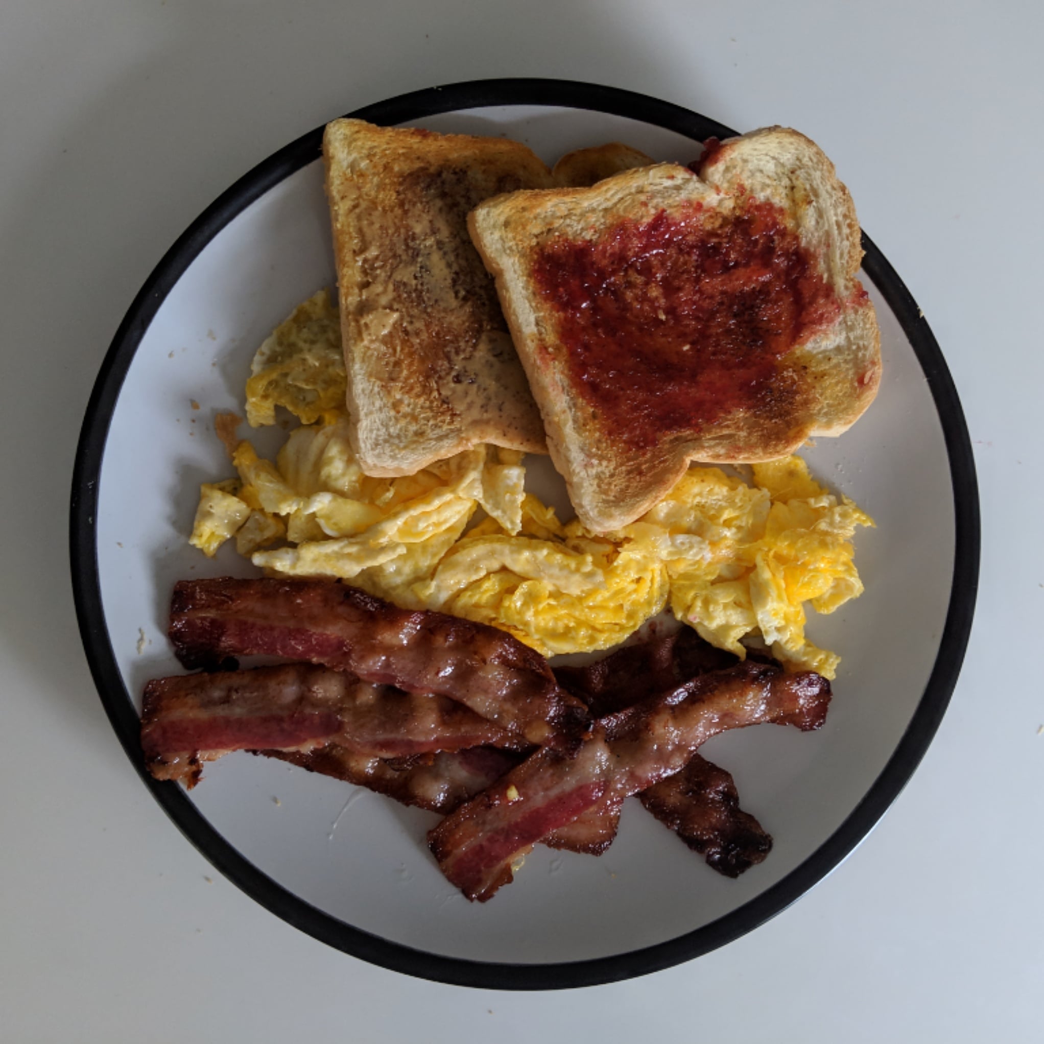 A plate with scrambled eggs, crispy bacon strips, and two slices of toast with red jelly on a white surface.