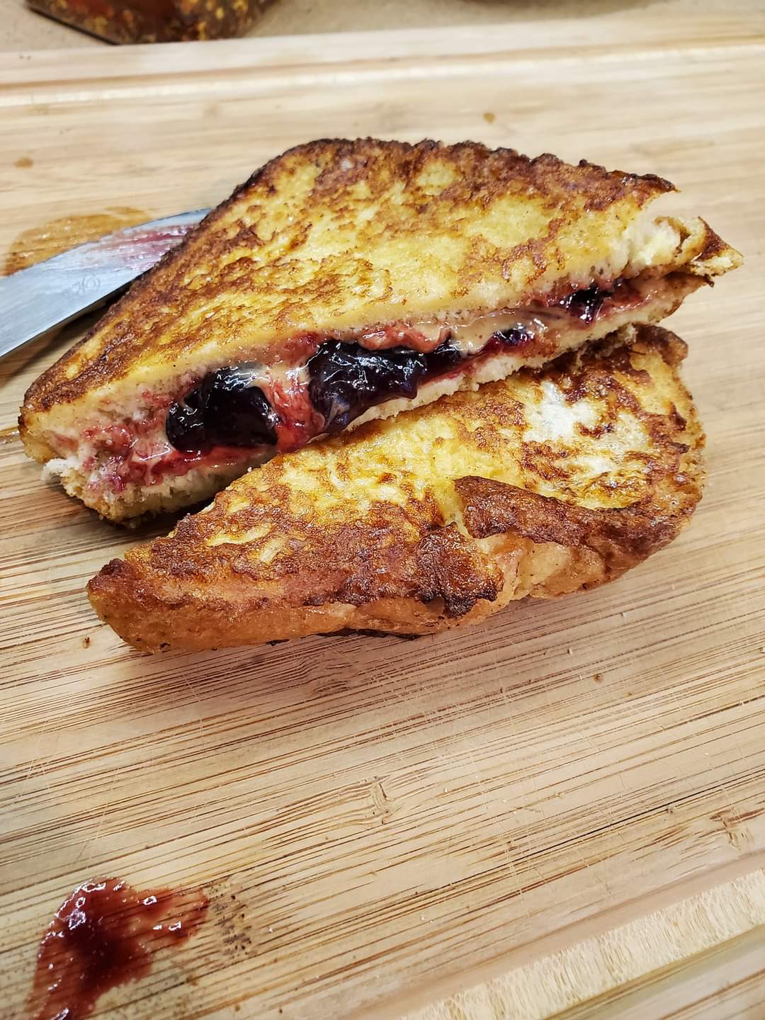 A grilled sandwich cut in half, filled with melted cheese and dark berry jam, sits on a wooden cutting board with a knife and a bit of jam spilling out.