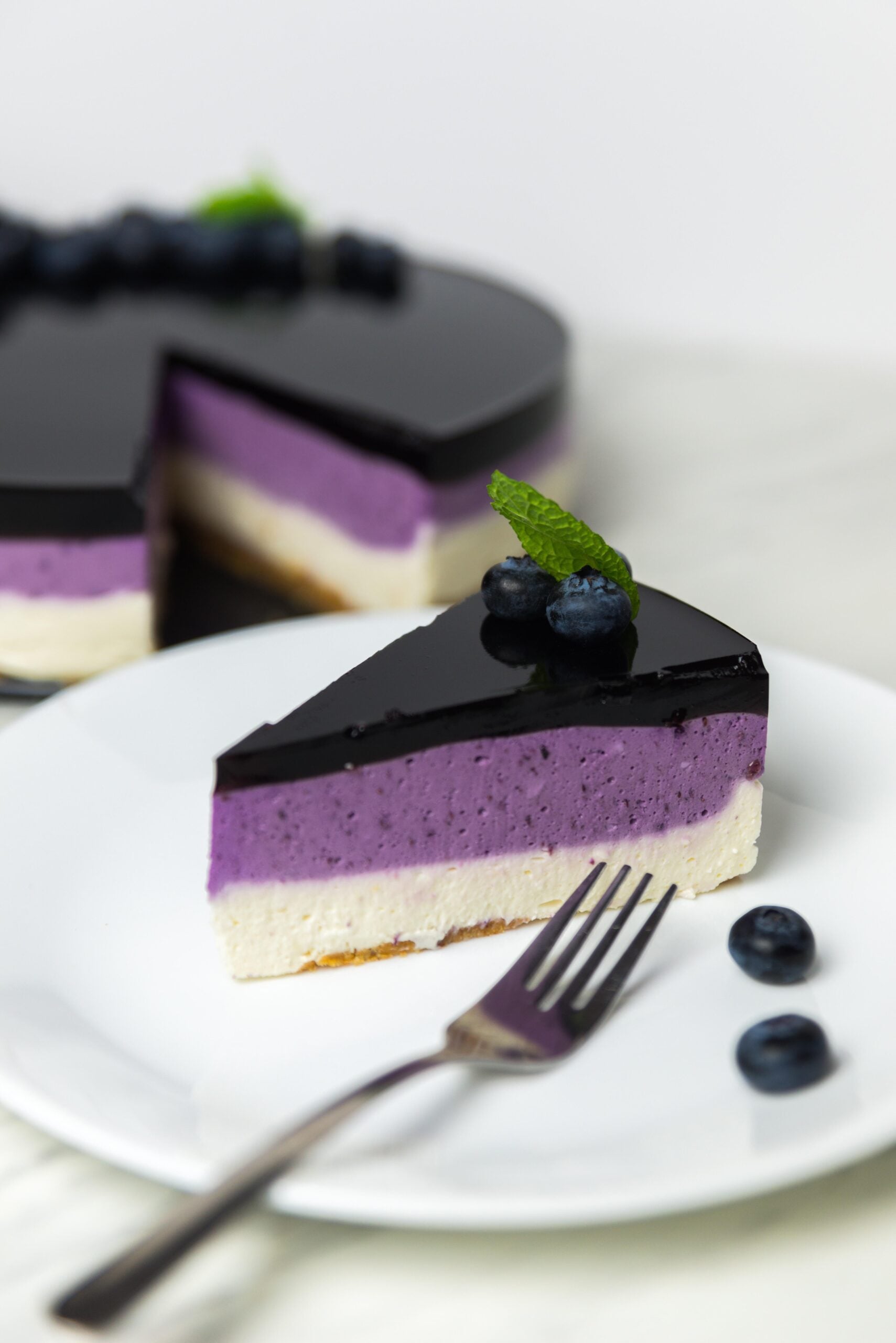 A slice of layered blueberry cheesecake on a white plate with blueberries and mint garnish, placed in front of the whole cake. A fork and extra blueberries are on the plate.