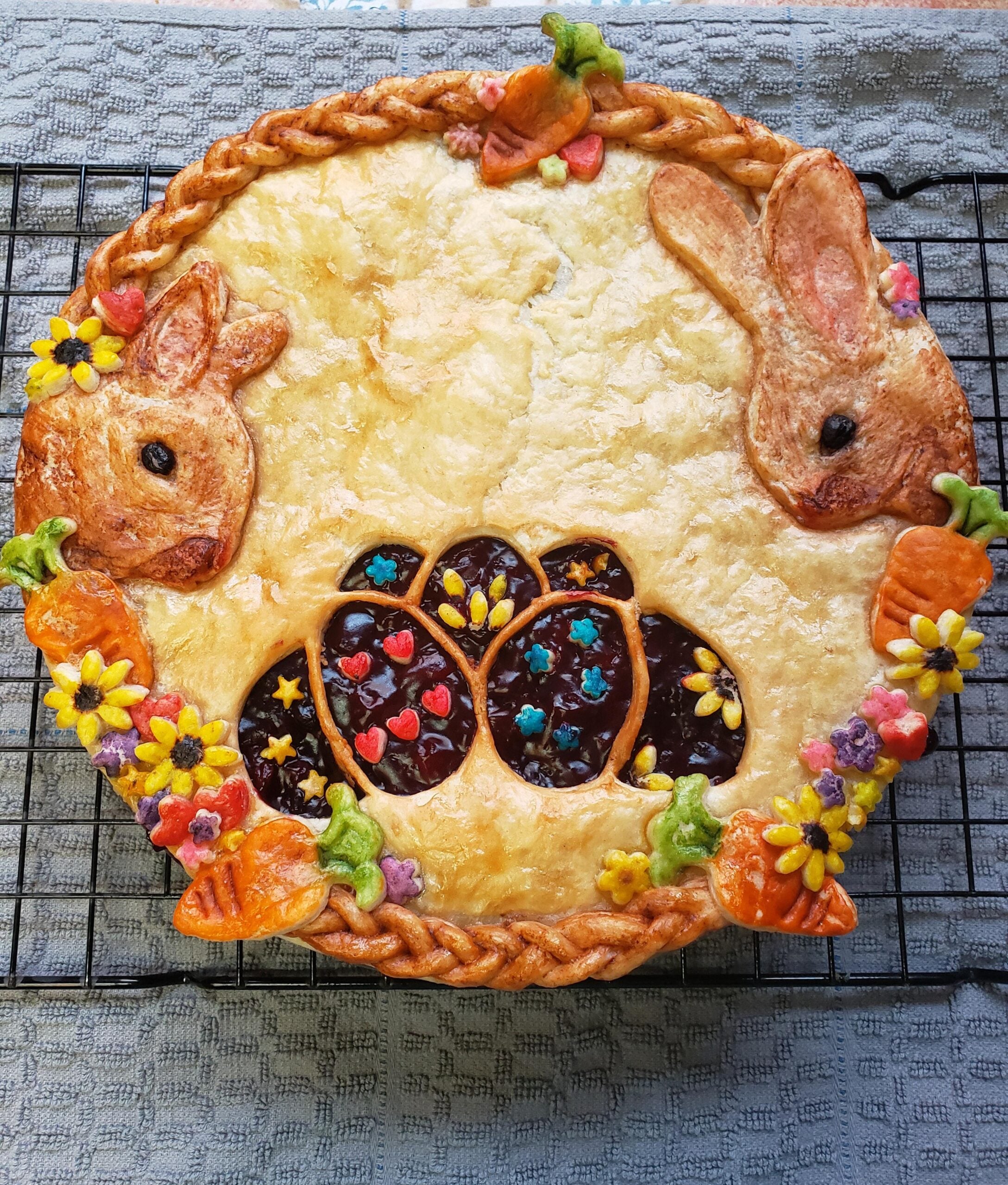 A pie with a golden crust decorated with pastry bunnies, carrots, colorful flowers, and three chocolate egg shapes in the center, viewed from above on a cooling rack.