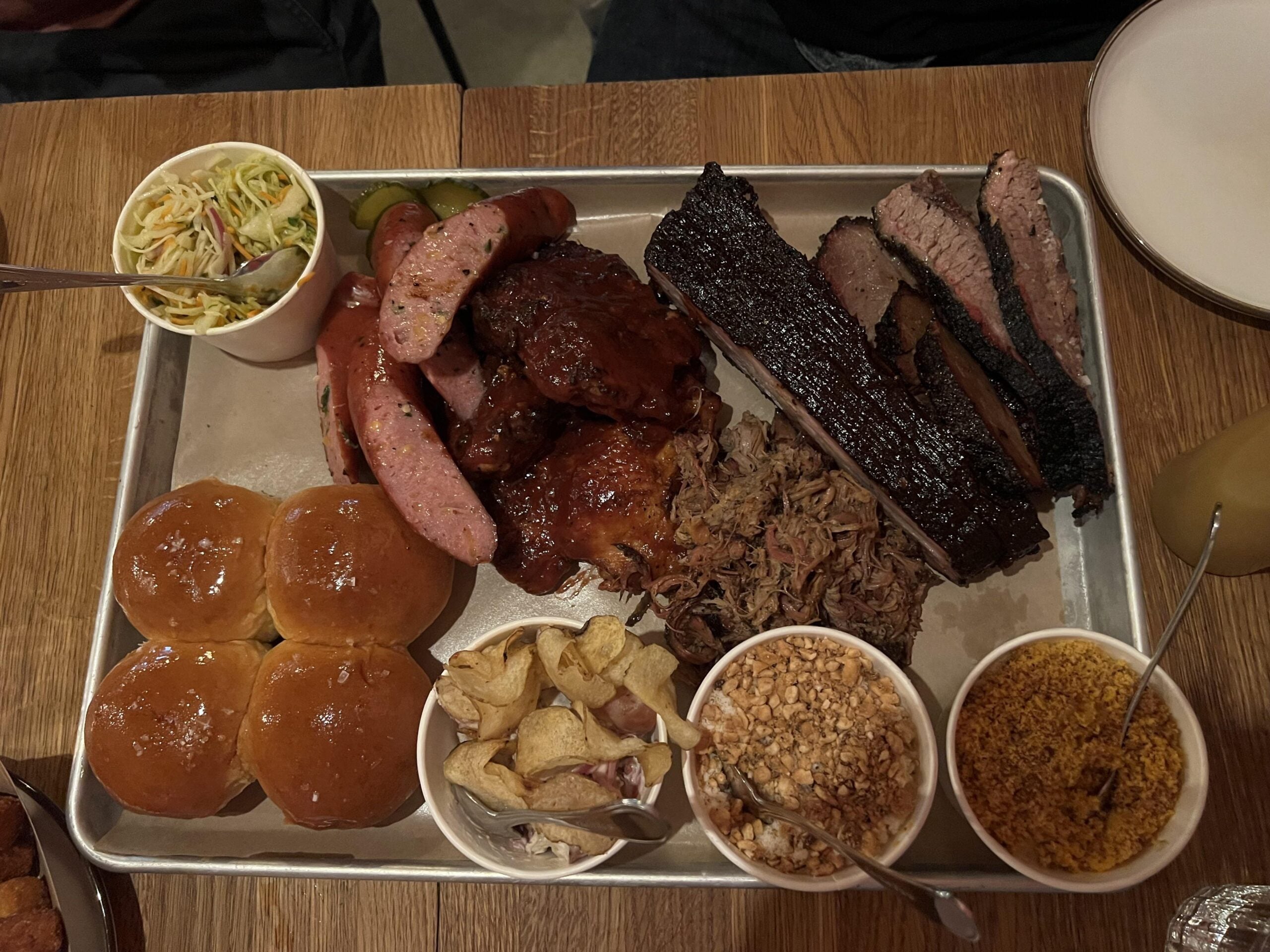 A tray of barbecue meats including brisket, ribs, sausage, pulled pork, and chicken—served with four rolls, coleslaw, potato chips, pickles, and two cups of baked beans and seasoned crumbs on a wooden table—makes for one of the best BBQ cookouts this summer.