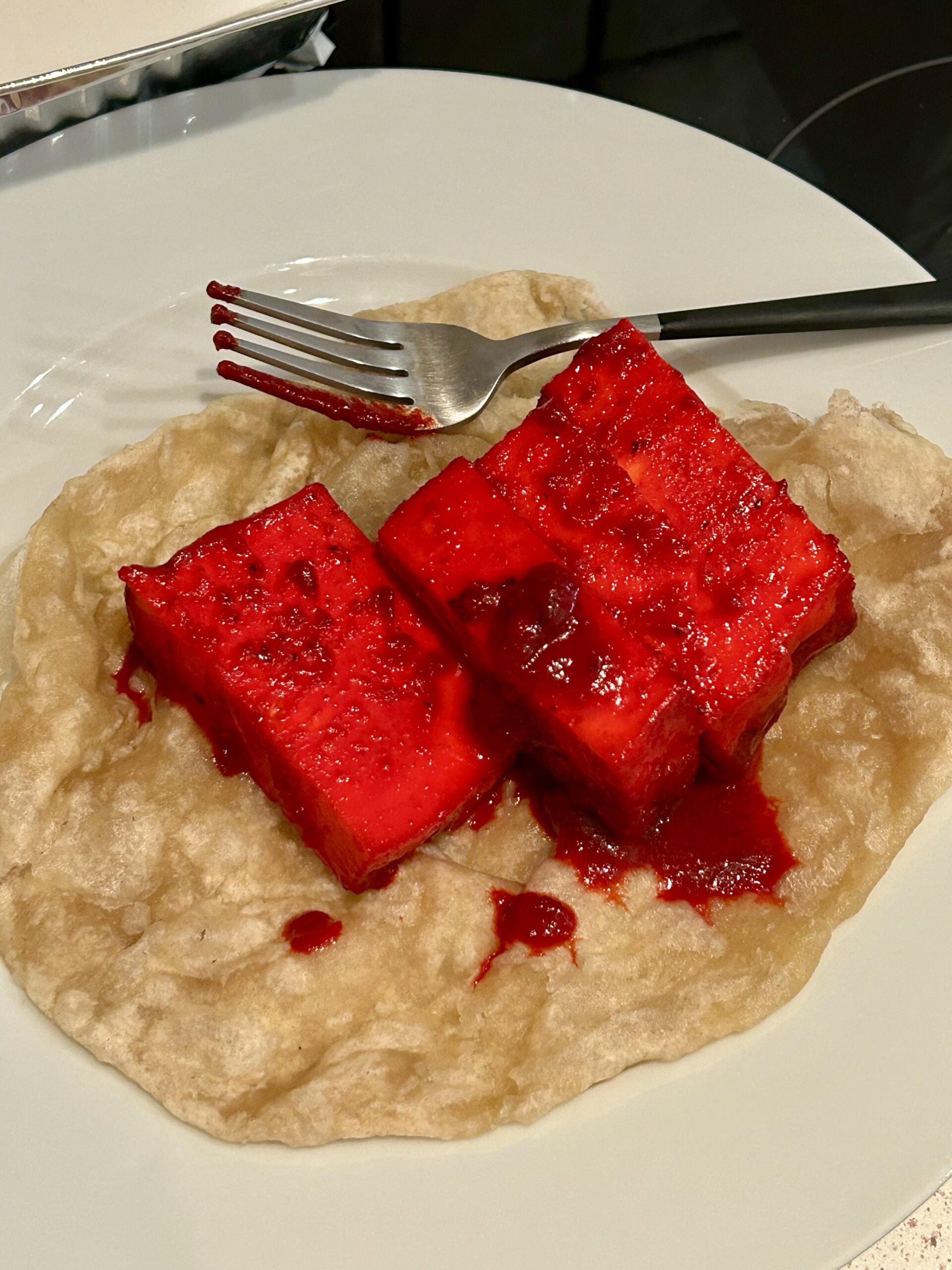 Two bright red cubes of watermelon with sauce sit on a light, puffy flatbread on a white plate, with a fork resting on top.