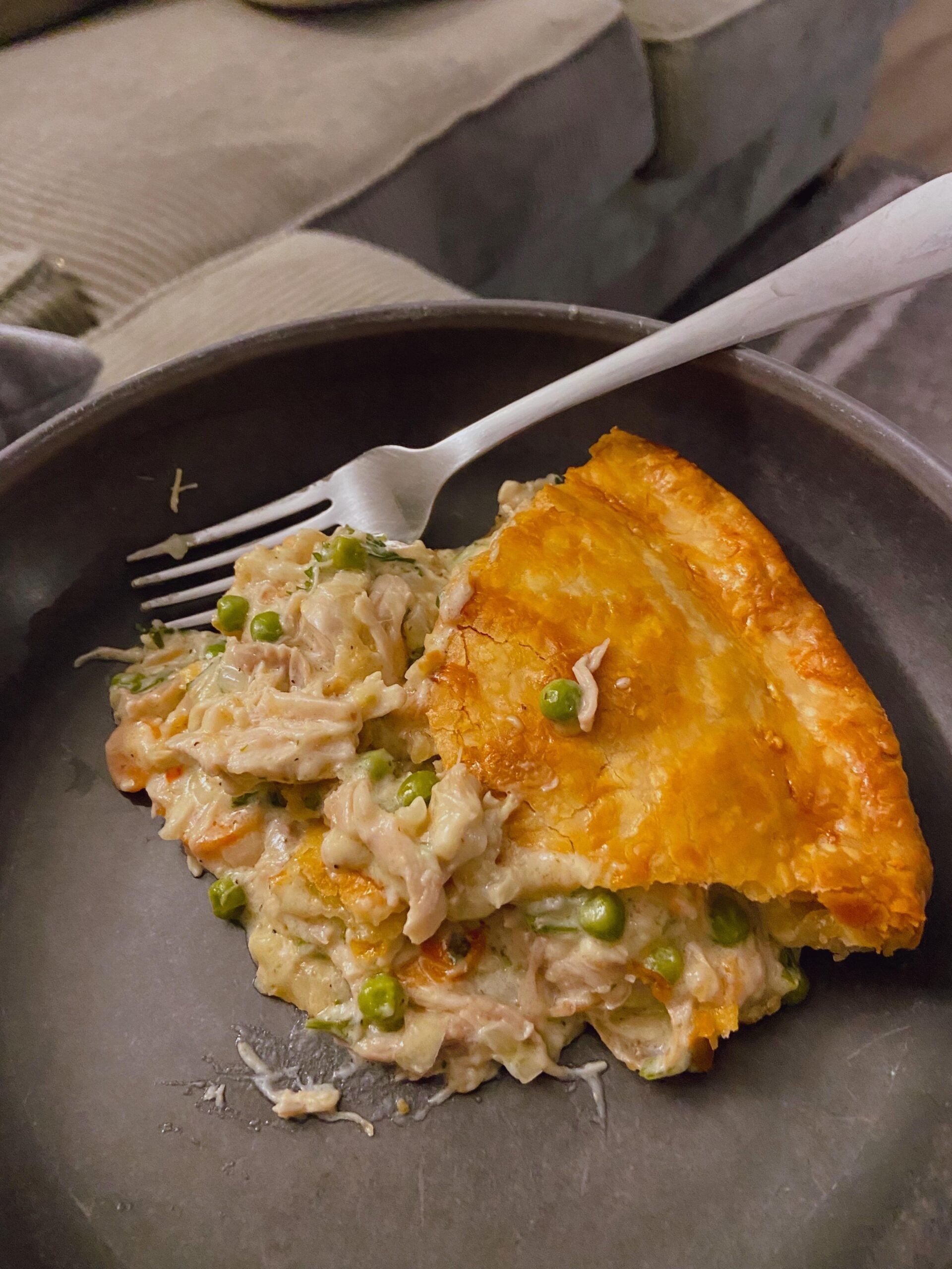 A slice of chicken pot pie with a golden crust, filled with shredded chicken, peas, and carrots, sits on a dark plate with a fork on a couch in the background.