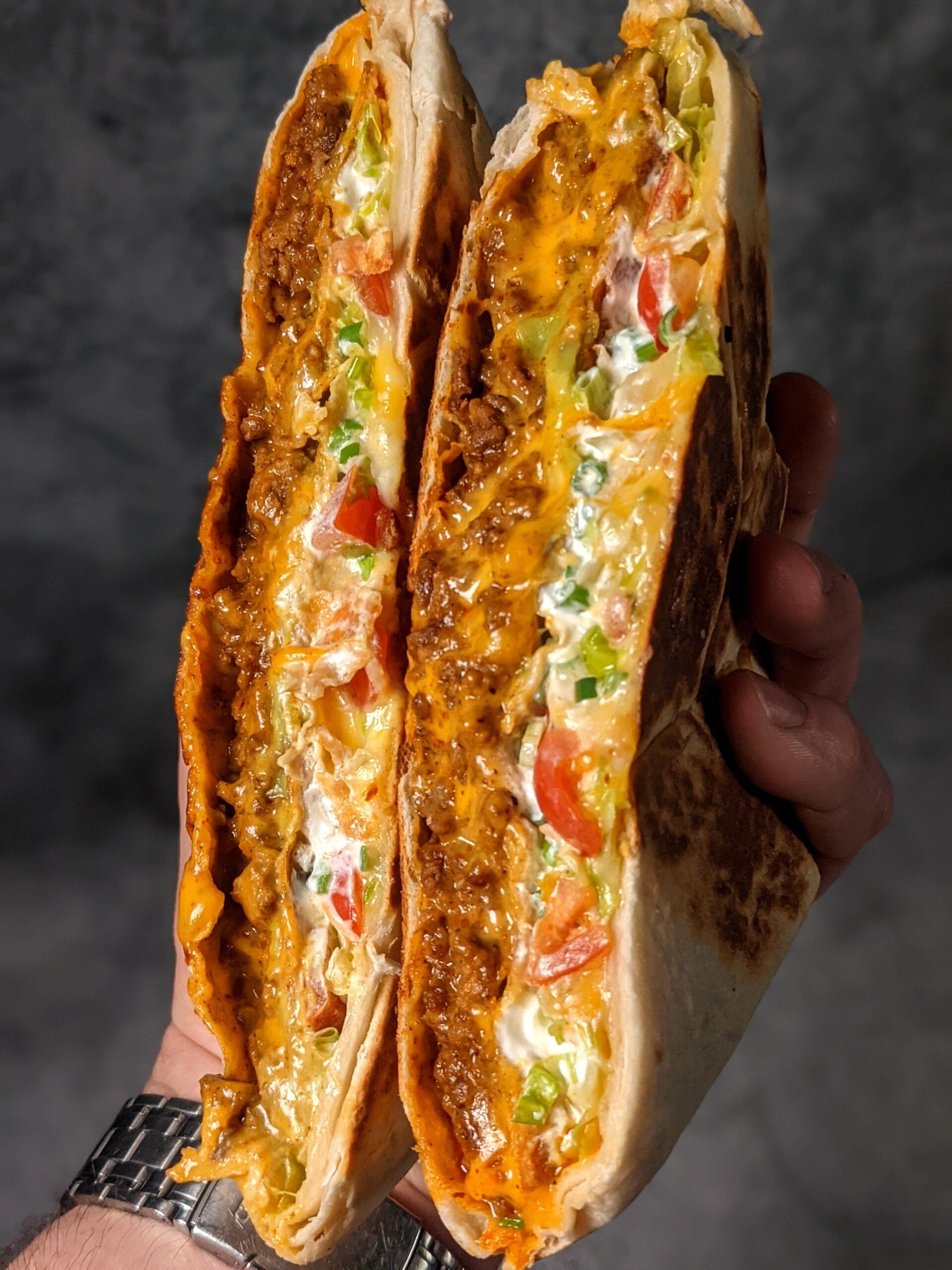 A hand holds a cut, grilled burrito filled with seasoned beef, melted cheese, lettuce, tomatoes, and creamy sauce, showing the colorful layers inside.