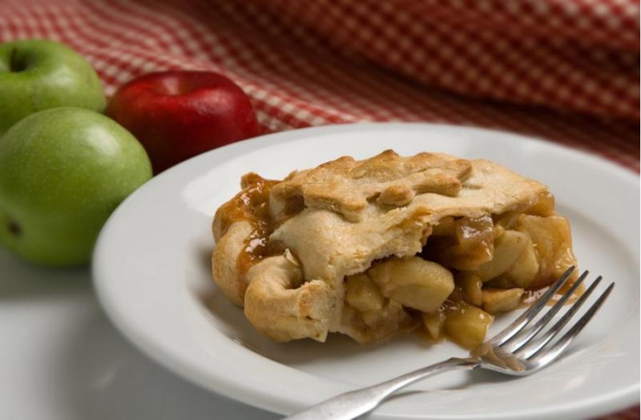 A slice of apple pie with a golden crust and visible apple filling sits on a white plate with a fork, perfectly capturing the essence of classic apple pies. In the background are two green apples, one red apple, and a red checkered cloth.