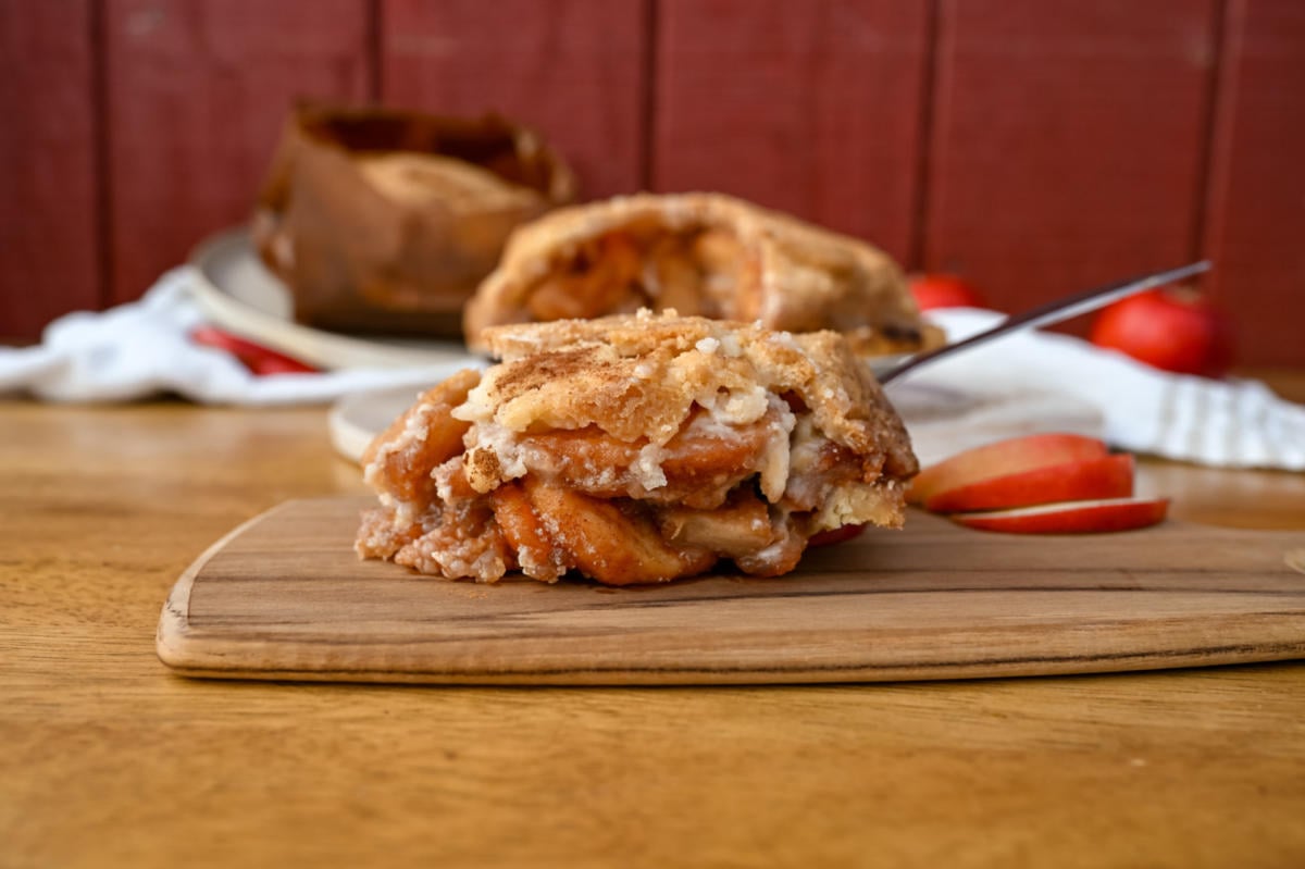 A slice of apple pie with a crumb topping sits on a wooden cutting board beside fresh apple slices. In the background, more apple pies and a red wooden wall are visible.