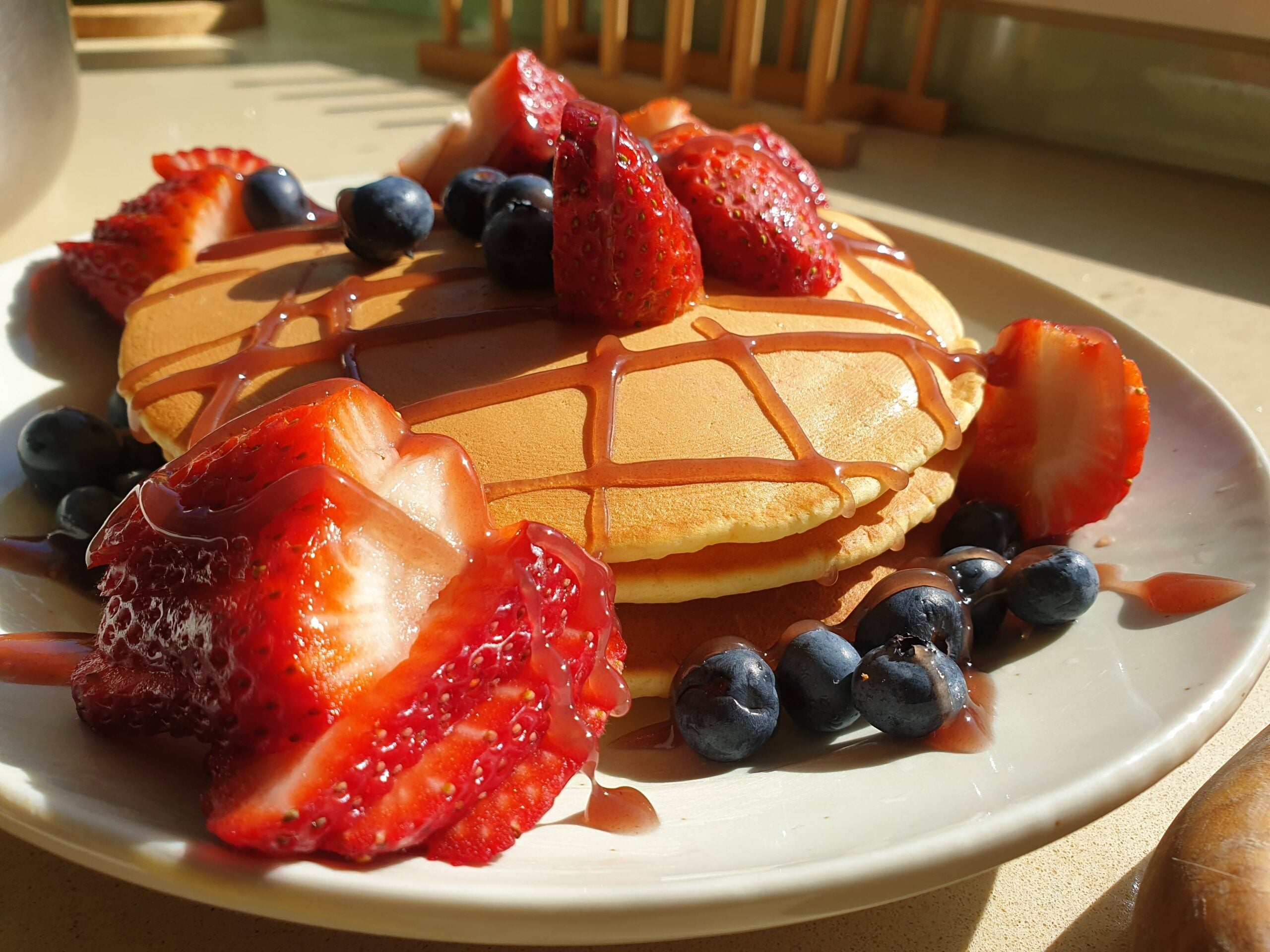 A plate of homemade pancakes topped with fresh strawberries, blueberries, and chocolate syrup, bathed in sunlight on a kitchen counter.