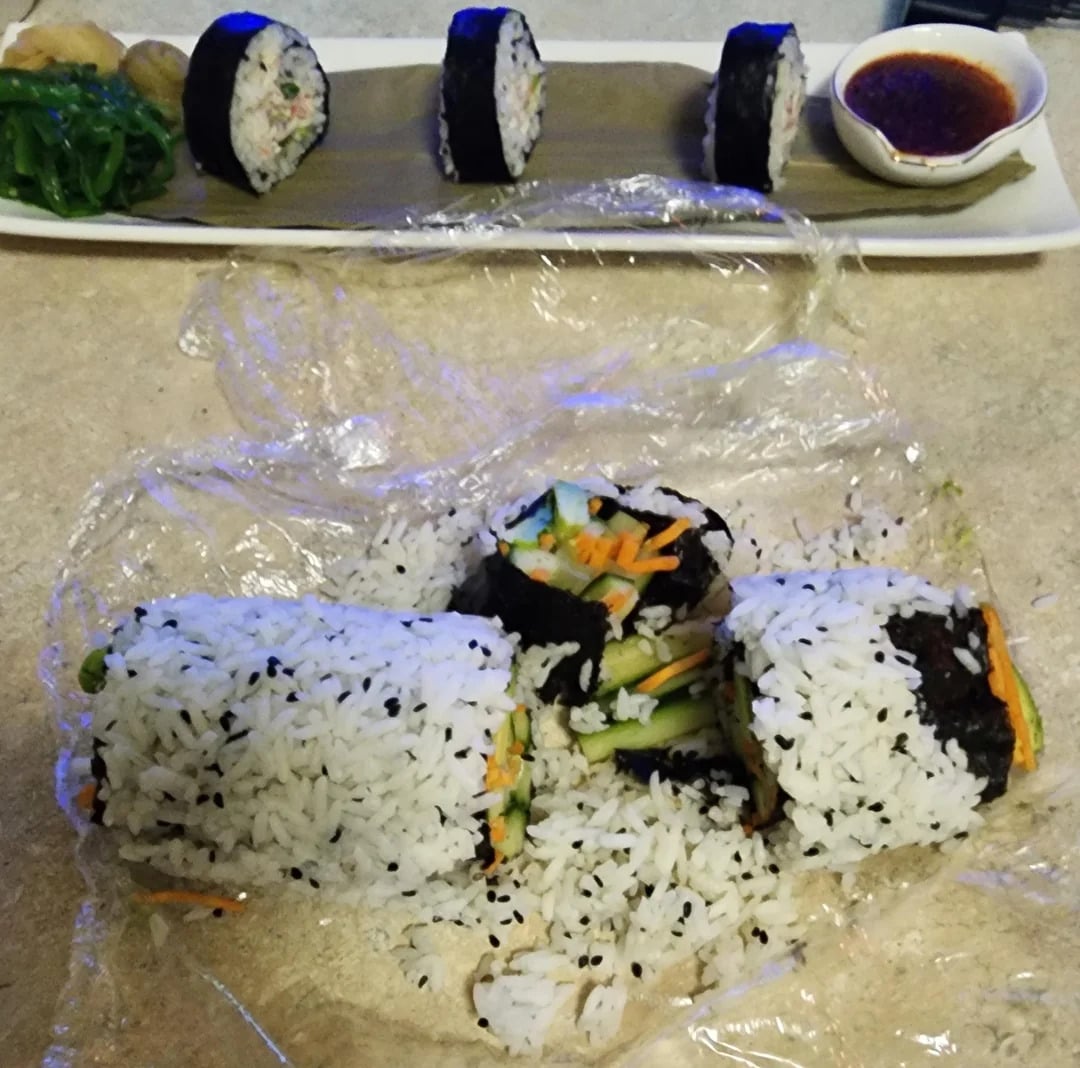 Two sushi rolls wrapped in plastic wrap on a table, with some pieces falling apart. In the background, neatly sliced sushi pieces are arranged on a rectangular plate with a small bowl of dipping sauce.