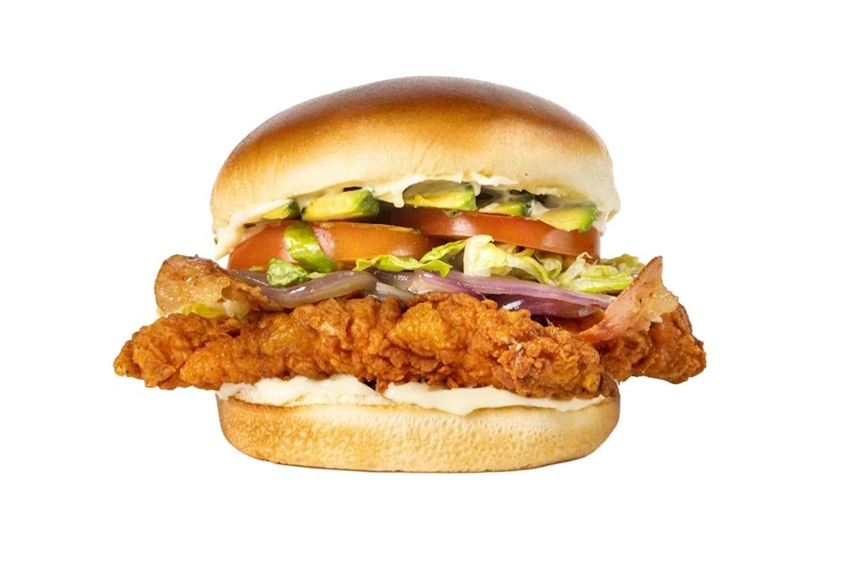 A crispy fried chicken sandwich—reminiscent of those served at popular fried chicken chains—with lettuce, tomato, pickles, onions, bacon, and mayonnaise on a toasted bun, shown against a white background.