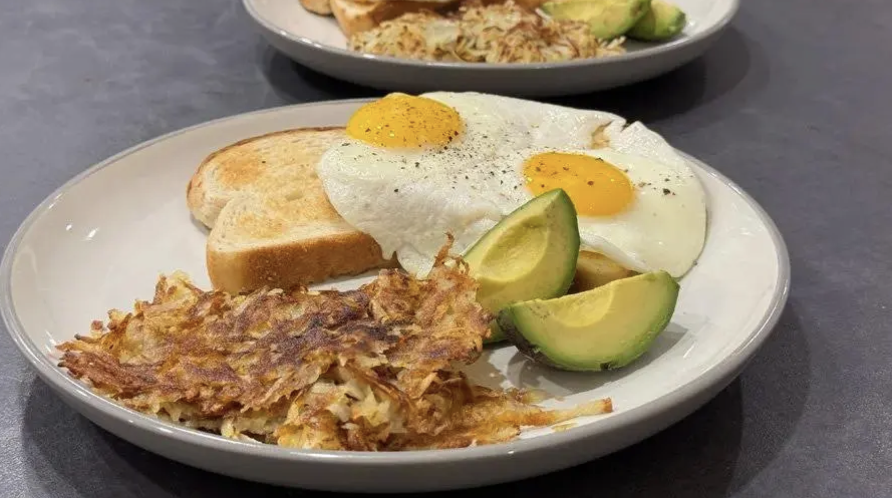 A breakfast plate with two sunny-side-up eggs on toast, sliced avocado, and a serving of crispy hash browns. Another similar plate is visible in the background.
