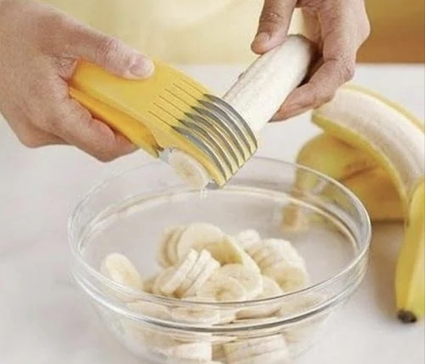 A person uses a yellow multi-blade slicer, one of the handy kitchen gadgets, to cut a banana into even slices over a glass bowl, with whole bananas in the background.