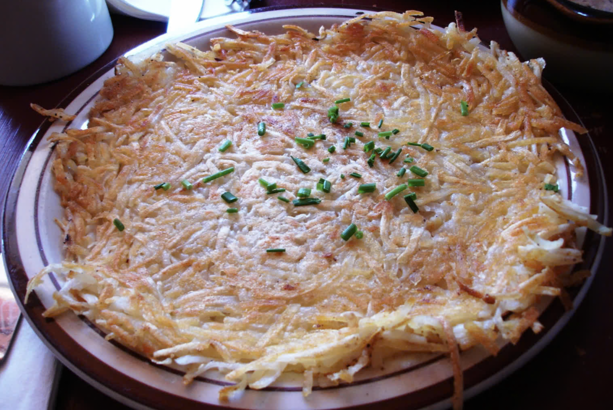 A round plate of crispy golden-brown hash browns, known as some of the best hash browns around, topped with chopped chives and served on a table.