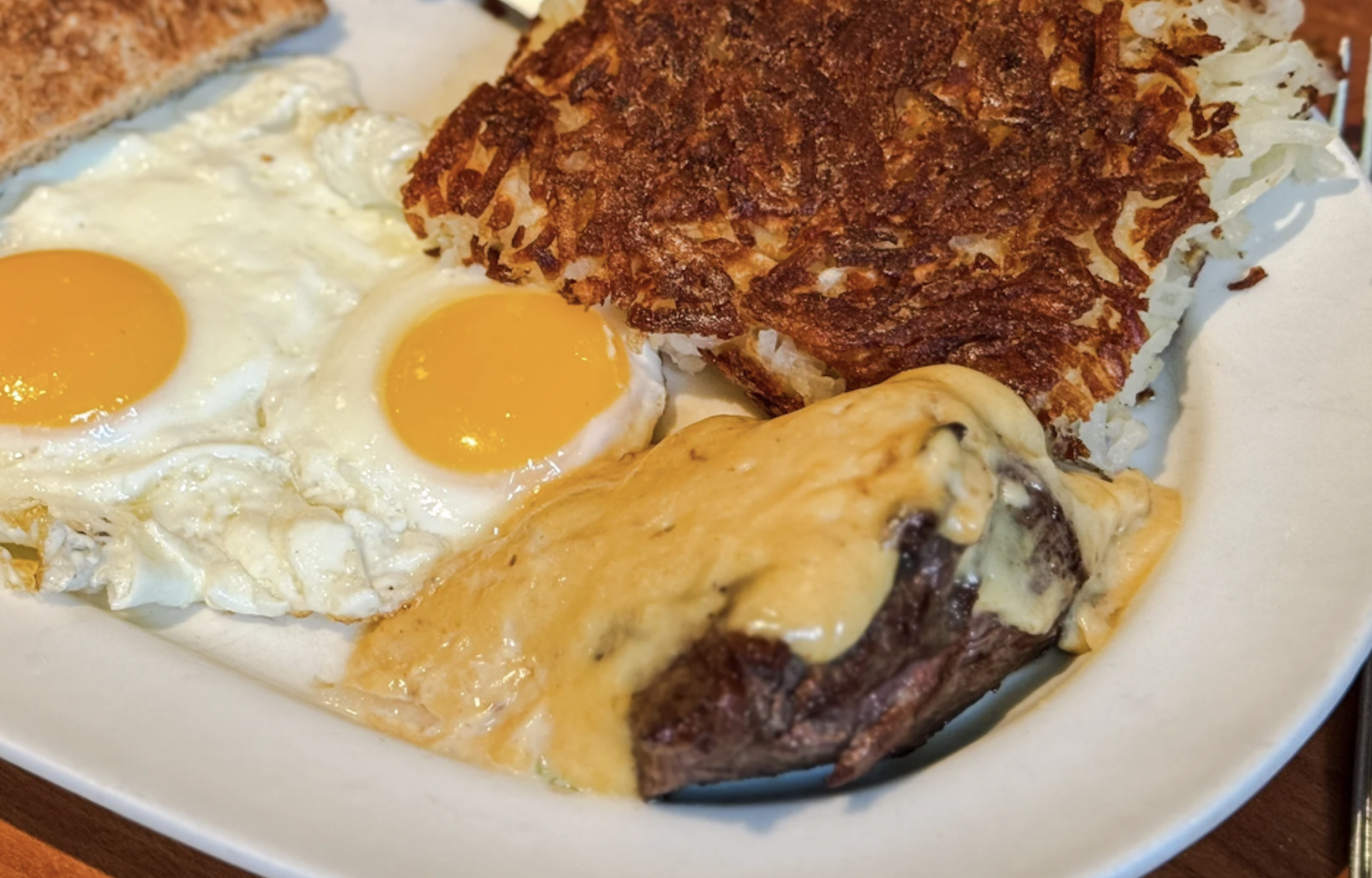 A breakfast plate with two sunny-side-up eggs, the best hash browns cooked to crispy perfection, a slice of toasted bread, and a steak topped with melted cheese sauce.