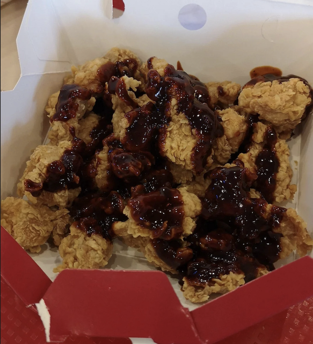 A box filled with golden fried chicken pieces topped with a thick, dark brown sauce, inside a red and white takeout container.