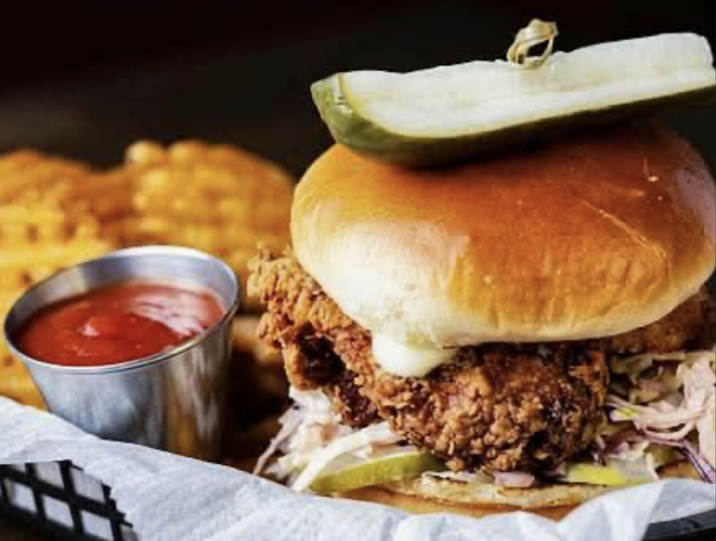 A fried chicken sandwich with coleslaw and pickles, topped with a pickle spear on a bun, served with waffle fries and a cup of ketchup.