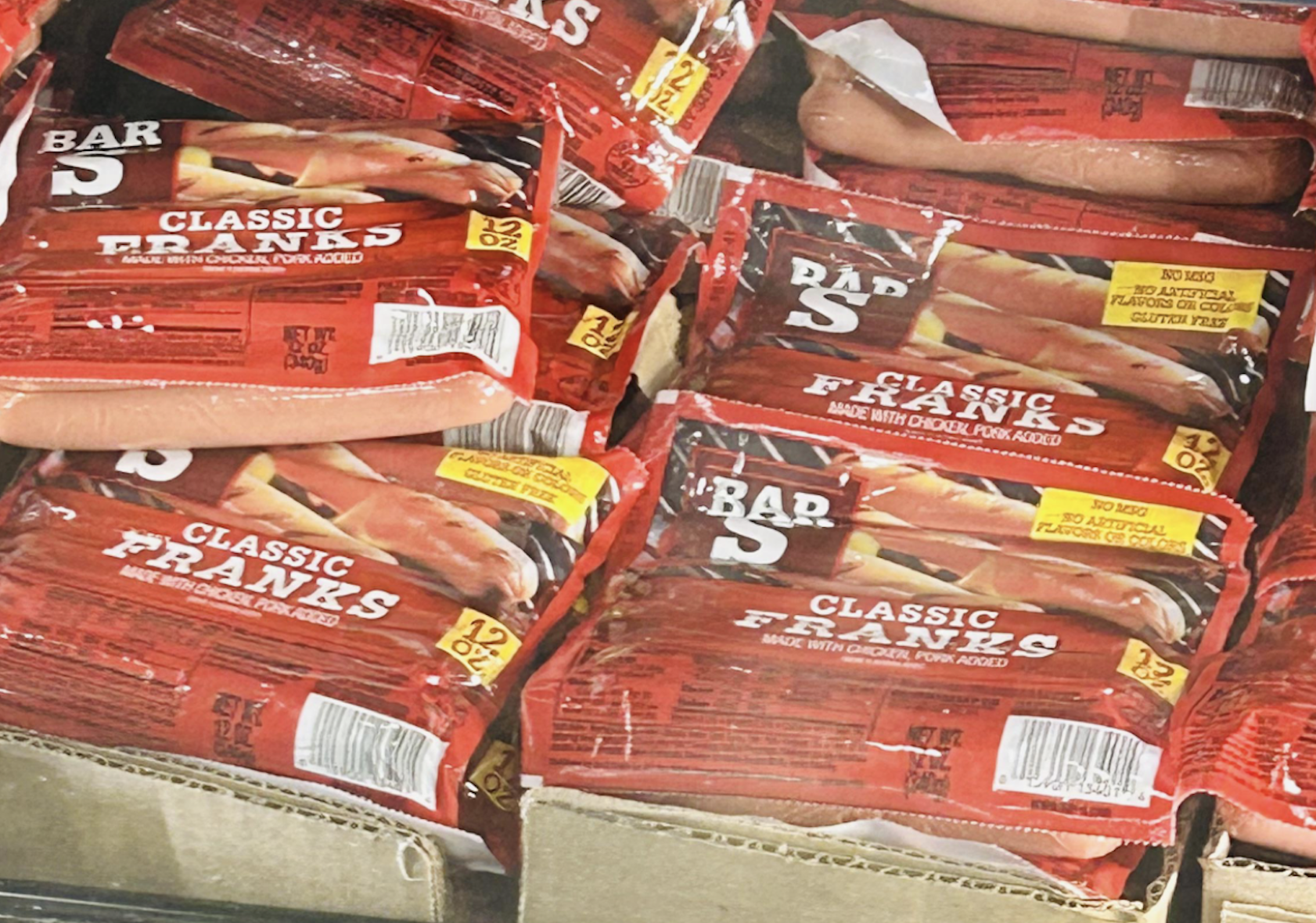 Several packages of Bar-S Classic Franks hot dogs are stacked in a cardboard box, with the red packaging and labels clearly visible. Some individual hot dogs are also visible in the upper left corner.