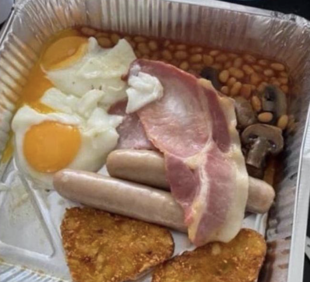 A takeaway tray with fried eggs, baked beans, mushrooms, two sausages, a slice of bacon, and two hash browns.