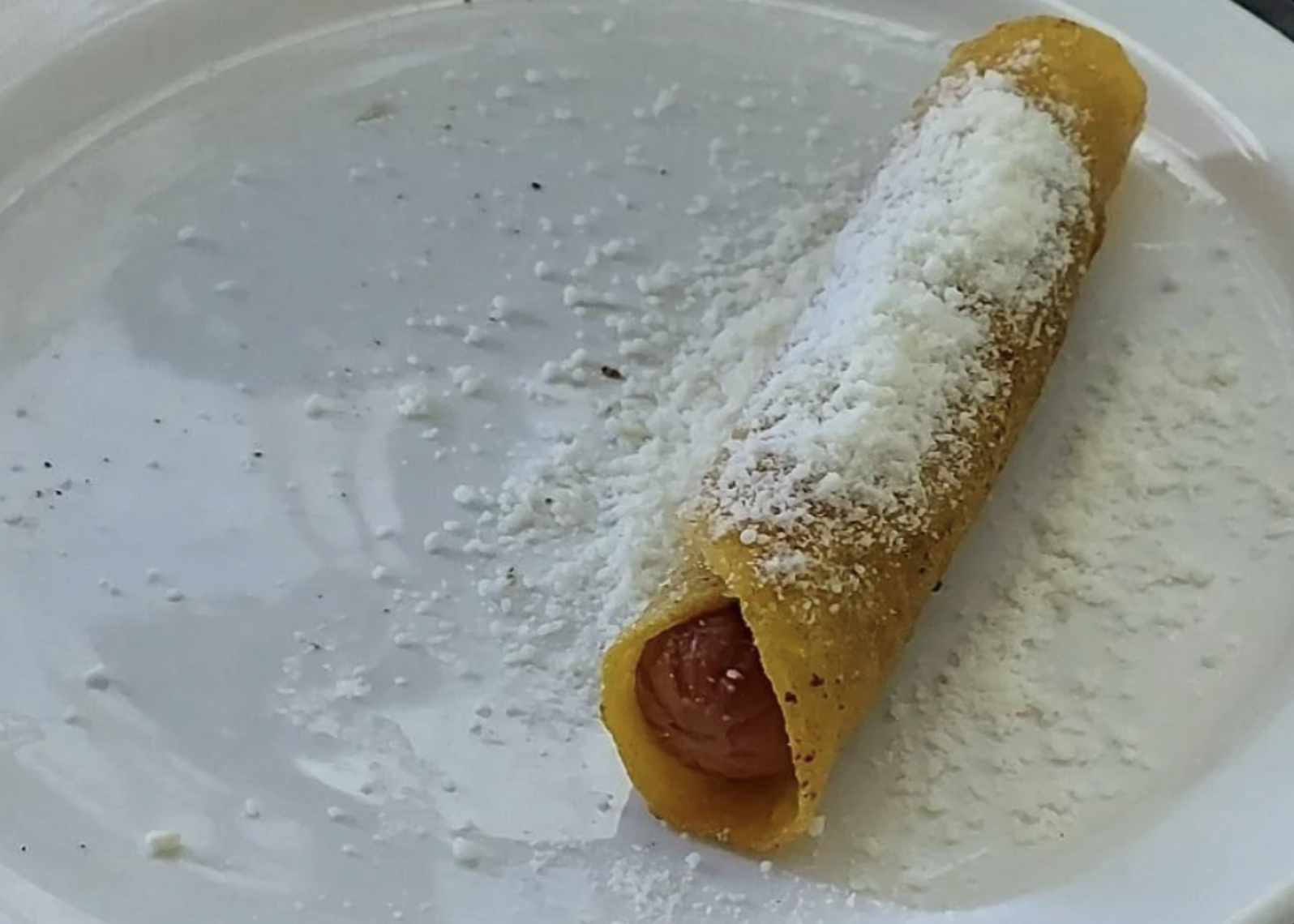 A hot dog is wrapped in a yellow corn tortilla, sprinkled with white grated cheese, and placed on a white plate with some cheese scattered around.