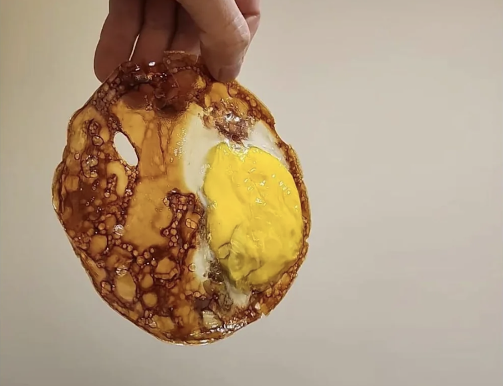 A hand holds up a thin, crispy, lacy fried egg with a yellow yolk and browned, irregularly shaped edges against a plain background.