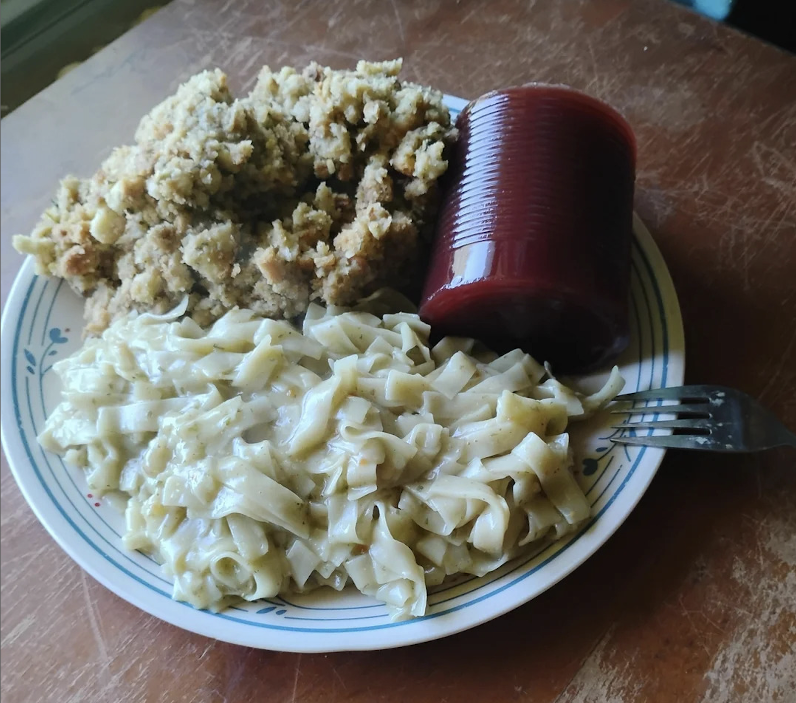 A plate with creamy noodles, a mound of stuffing, and a sliced cylinder of canned cranberry sauce on a wooden table, with a fork resting on the plate.