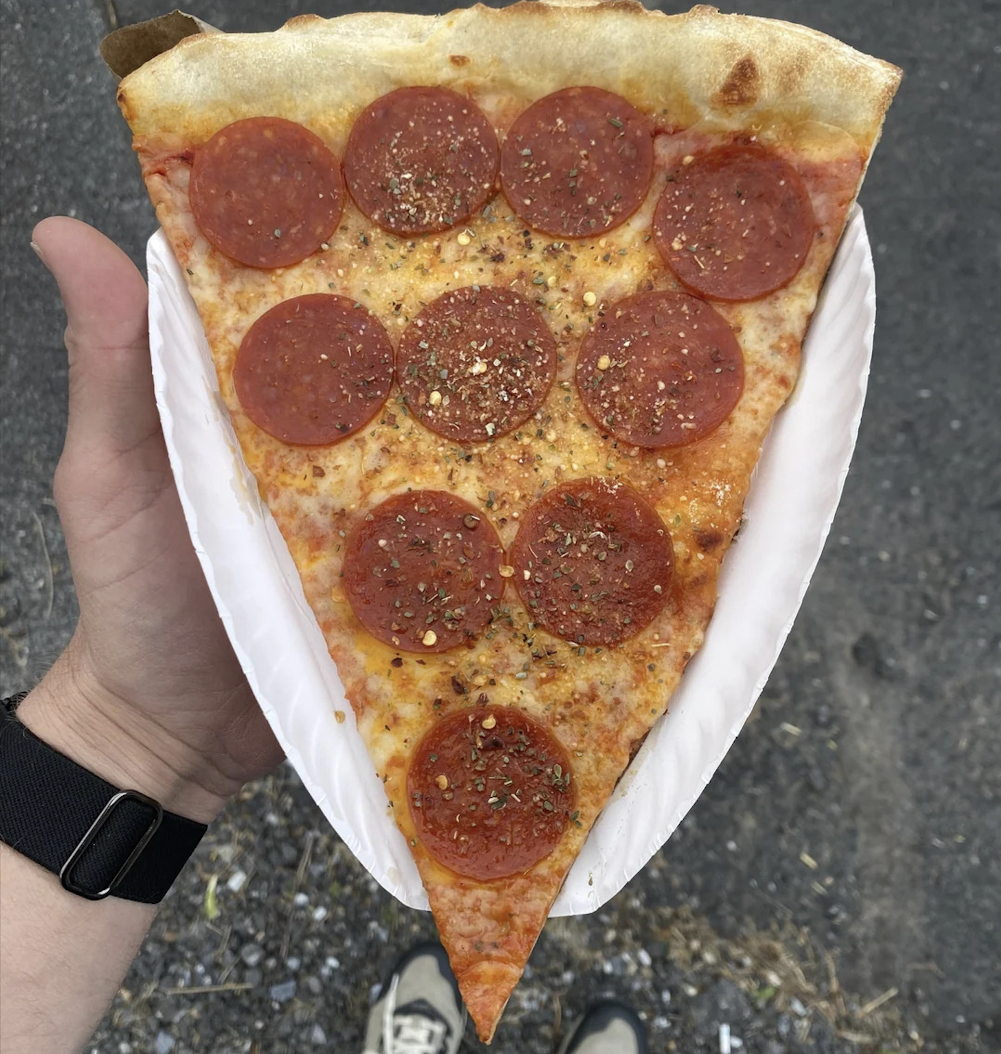 A hand holding a large slice of pepperoni pizza on a white paper plate, topped with pepper flakes—one of the best pizza photos for capturing that perfect outdoor pavement moment.