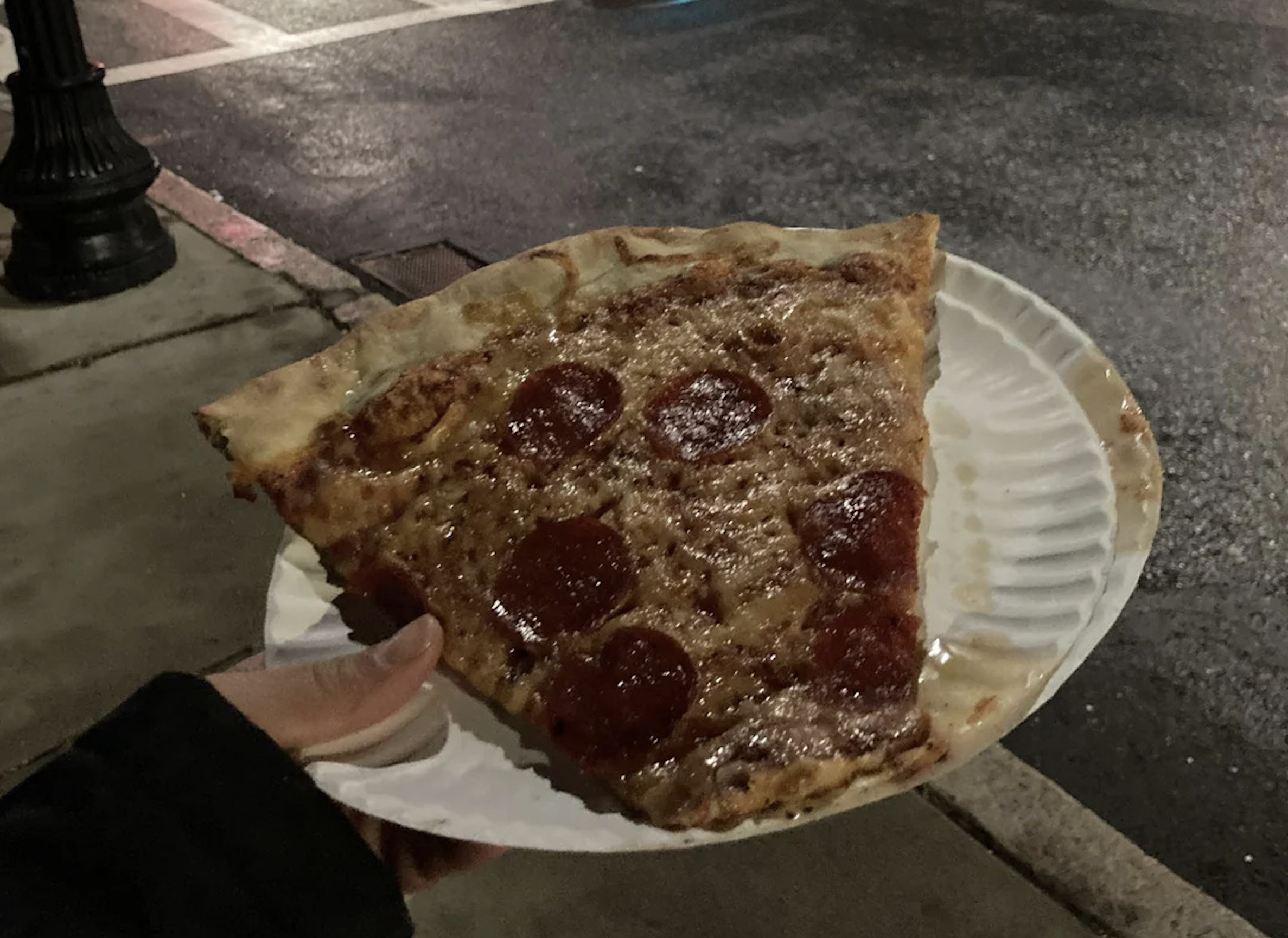 A hand holds a large slice of pepperoni pizza on a white paper plate, outdoors at night near a wet street and sidewalk—capturing one of the best pizza photos with its mouthwatering detail and urban atmosphere.