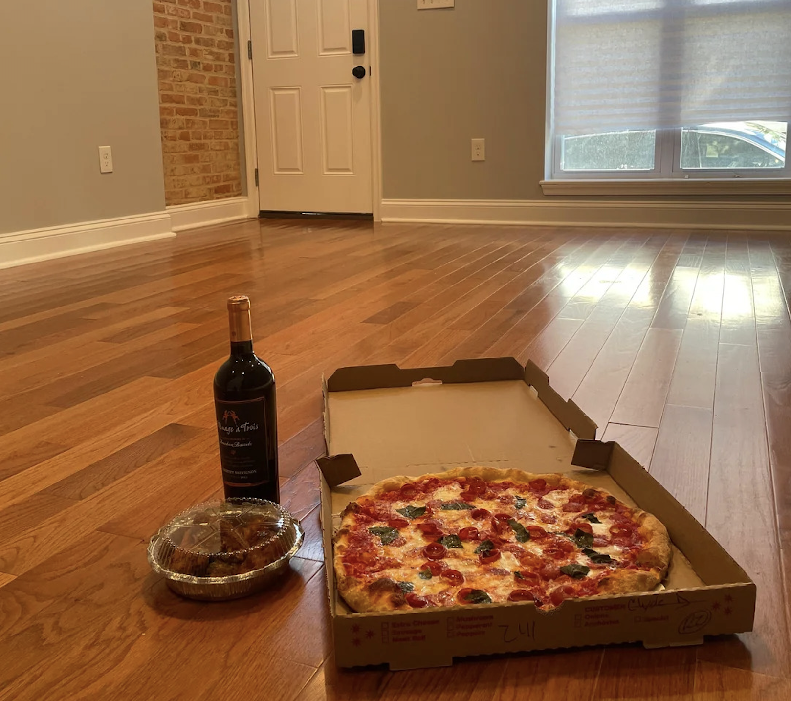 A pizza in an open box, a bottle of wine, and a container of food sit on a hardwood floor in an empty room with sunlight coming through a window—capturing one of the best pizza photos for those who appreciate simple pleasures.