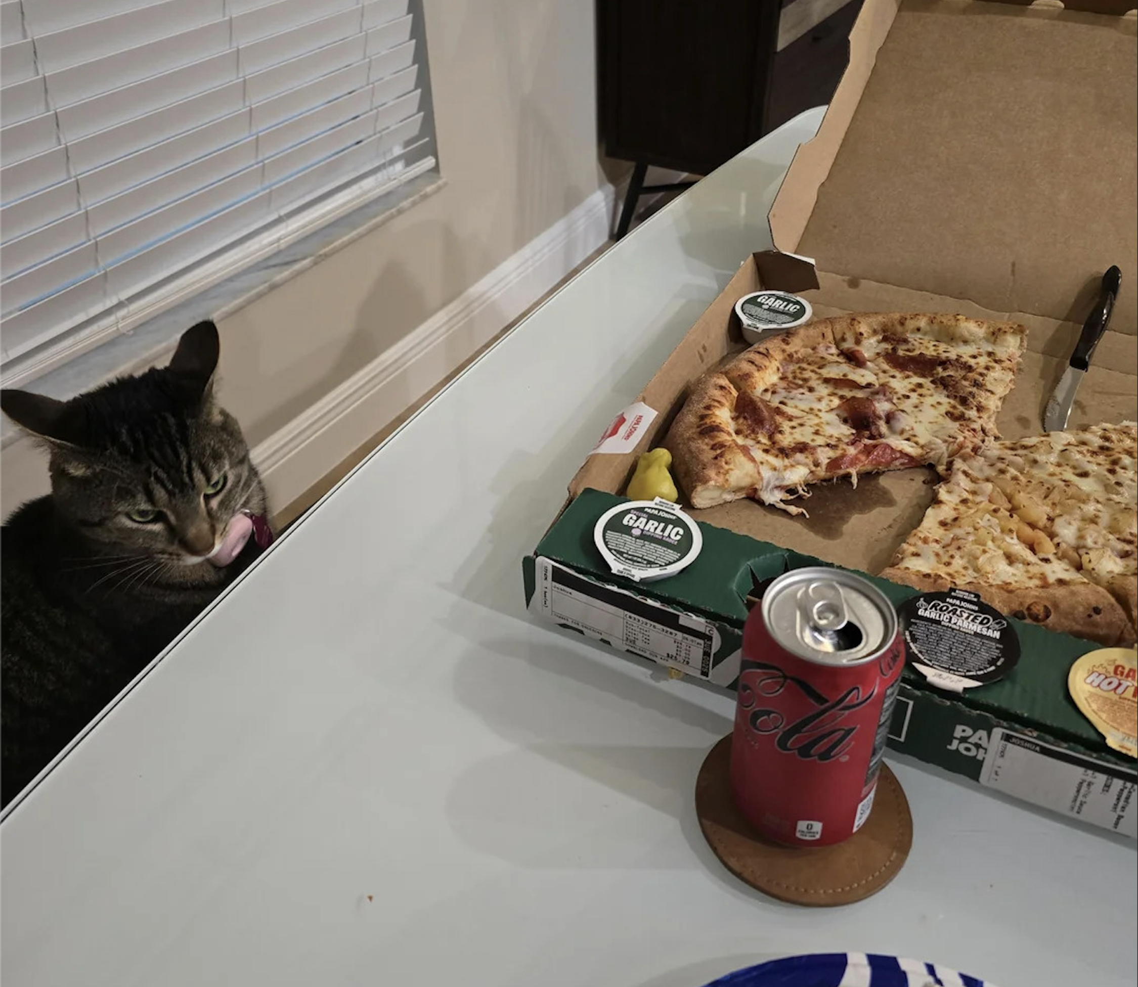 A tabby cat licks its lips near a table with an open pizza box, garlic sauce, and a can of Coca-Cola. The pizza is partly eaten, and the setting appears to be a casual dining area—capturing one of the best pizza photos for anyone who loves sharing quirky food moments with their pets.