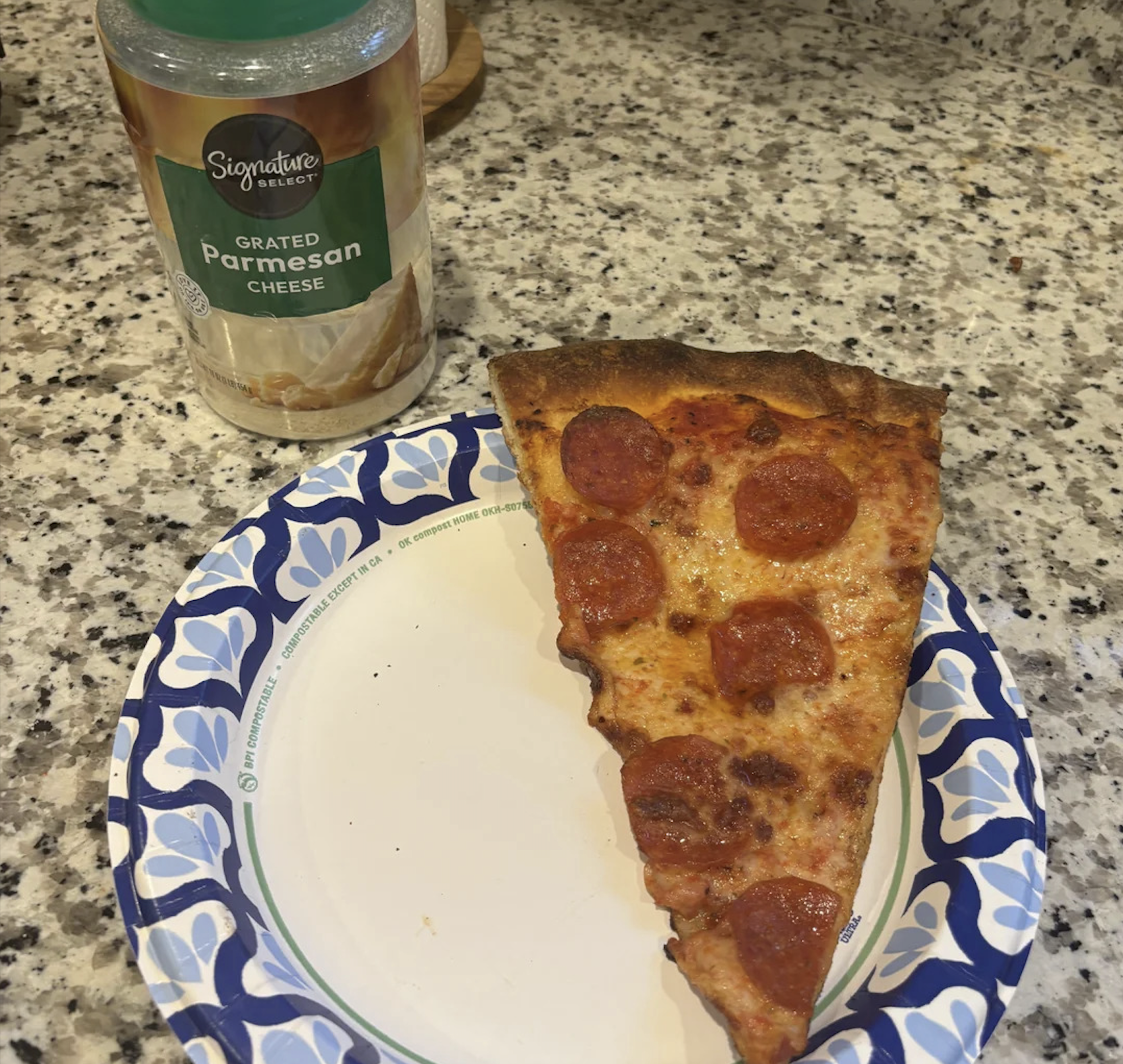 A slice of pepperoni pizza sits on a paper plate beside a container of grated parmesan cheese on a granite countertop, capturing one of the best pizza photos for any food lover.