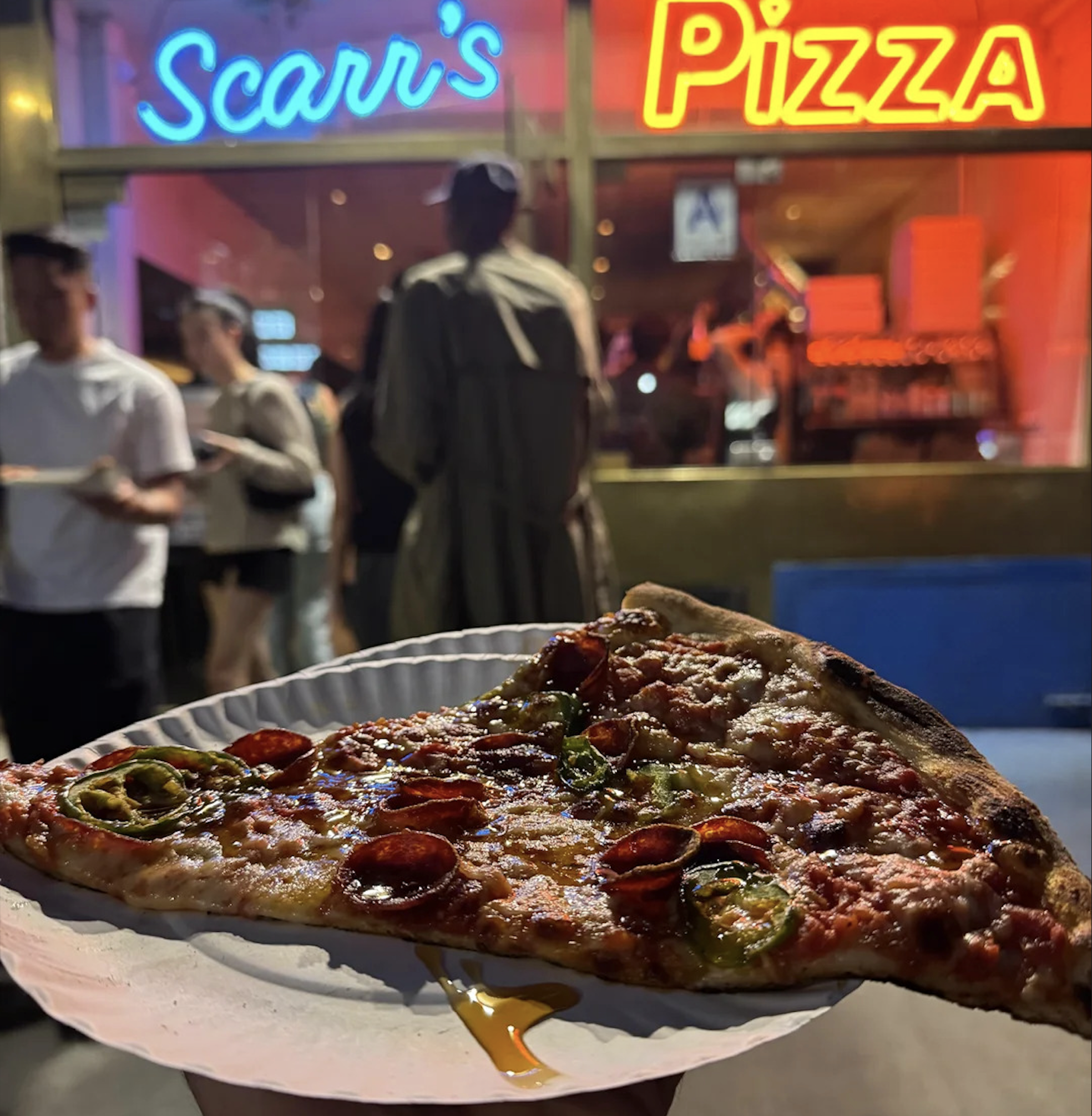 A close-up of a slice of pepperoni pizza on a paper plate is held outside Scarr's Pizza shop at night, with neon signs and people in the background—capturing one of the best pizza photos for any true food lover.