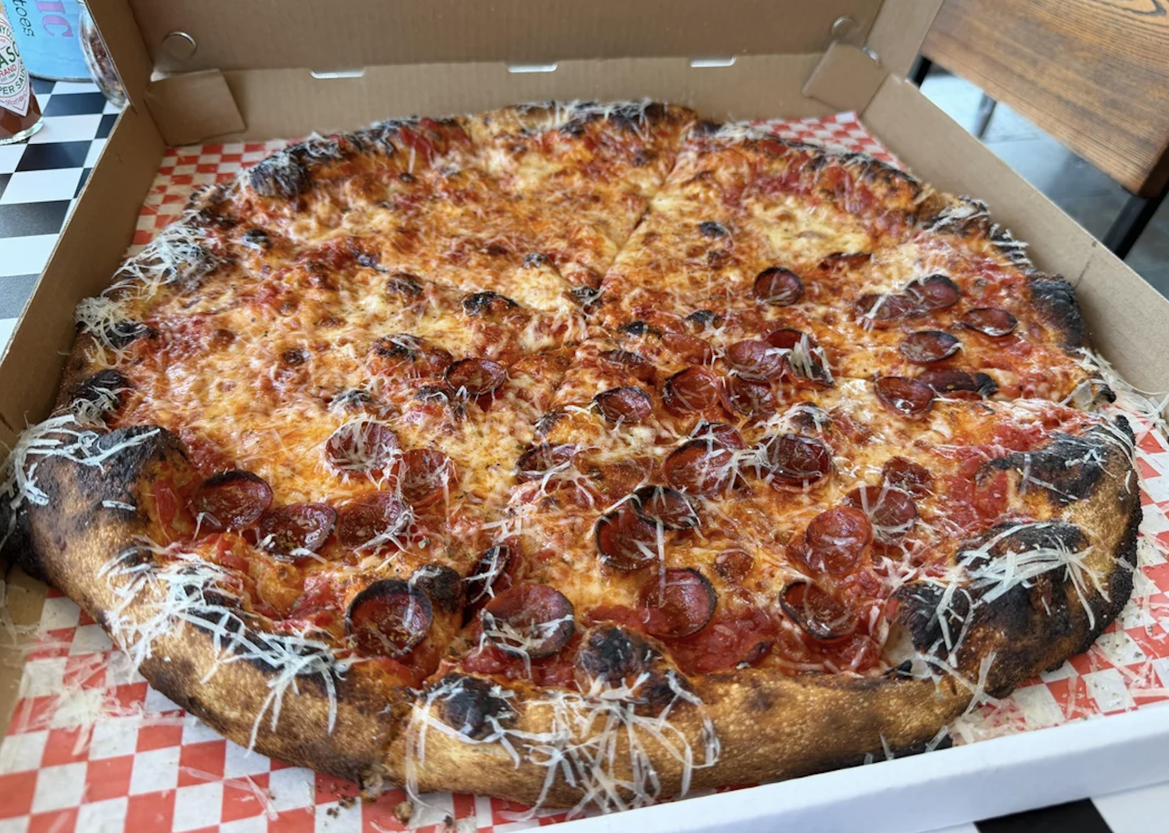 A pepperoni pizza with a crispy, slightly burnt crust sits in an open pizza box. The pizza is topped with melted cheese, sliced pepperoni, and grated parmesan on a red and white checkered liner. Captured as one of the best pizza photos, this image perfectly showcases the irresistible combination of flavors and textures.