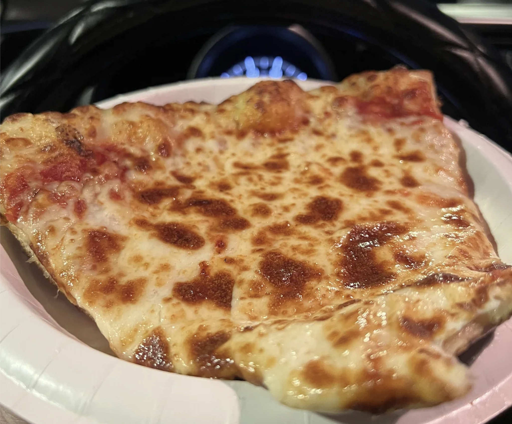 A close-up of a slice of cheese pizza with a golden, bubbly crust, sitting on a white paper plate inside a car—one of the best pizza photos capturing that perfect cheesy moment.