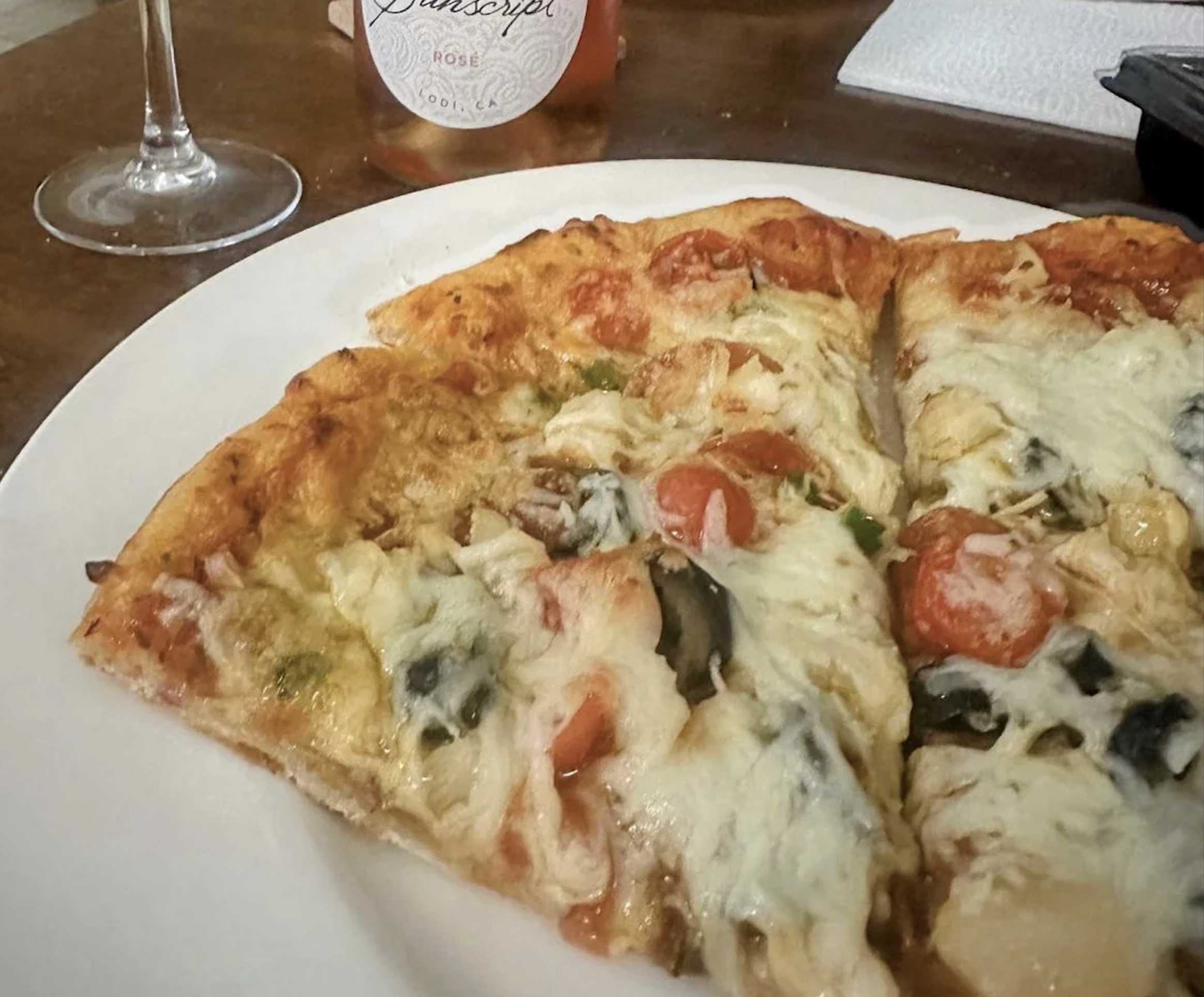 A close-up of three slices of veggie pizza with melted cheese, tomatoes, and spinach on a white plate showcases one of the best pizza photos. A wine glass and a bottle of rosé wine are visible in the background.