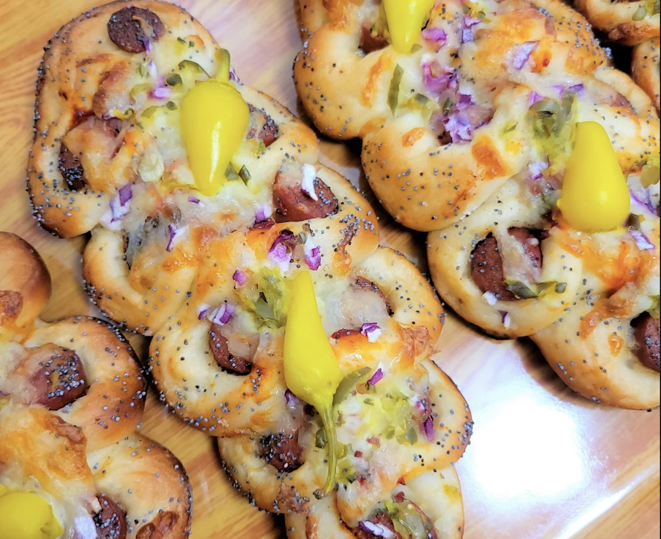 Braided bread loaves topped with melted cheese, sliced sausage, chopped red onions, jalapeƱos, and a yellow pepper, served on a wooden tray.