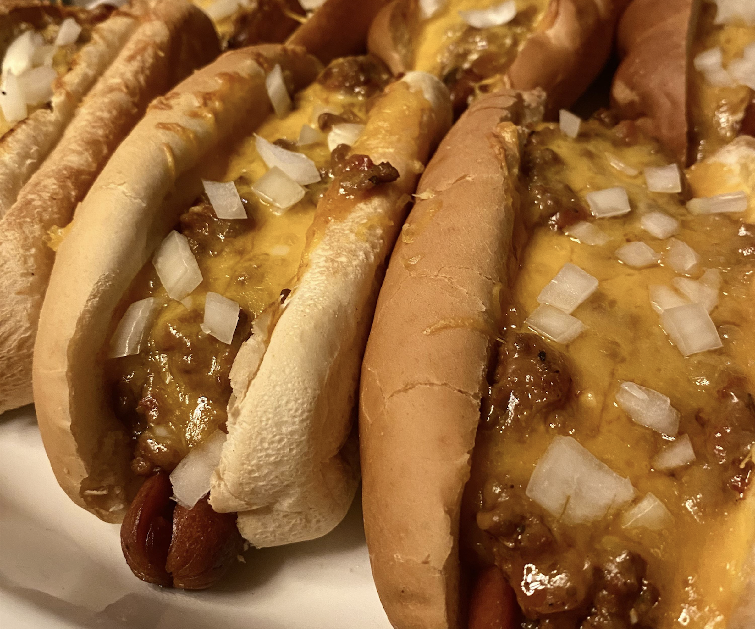 Close-up of several chili cheese hot dogs in buns, topped with melted cheddar cheese and chopped onions, arranged on a plate.
