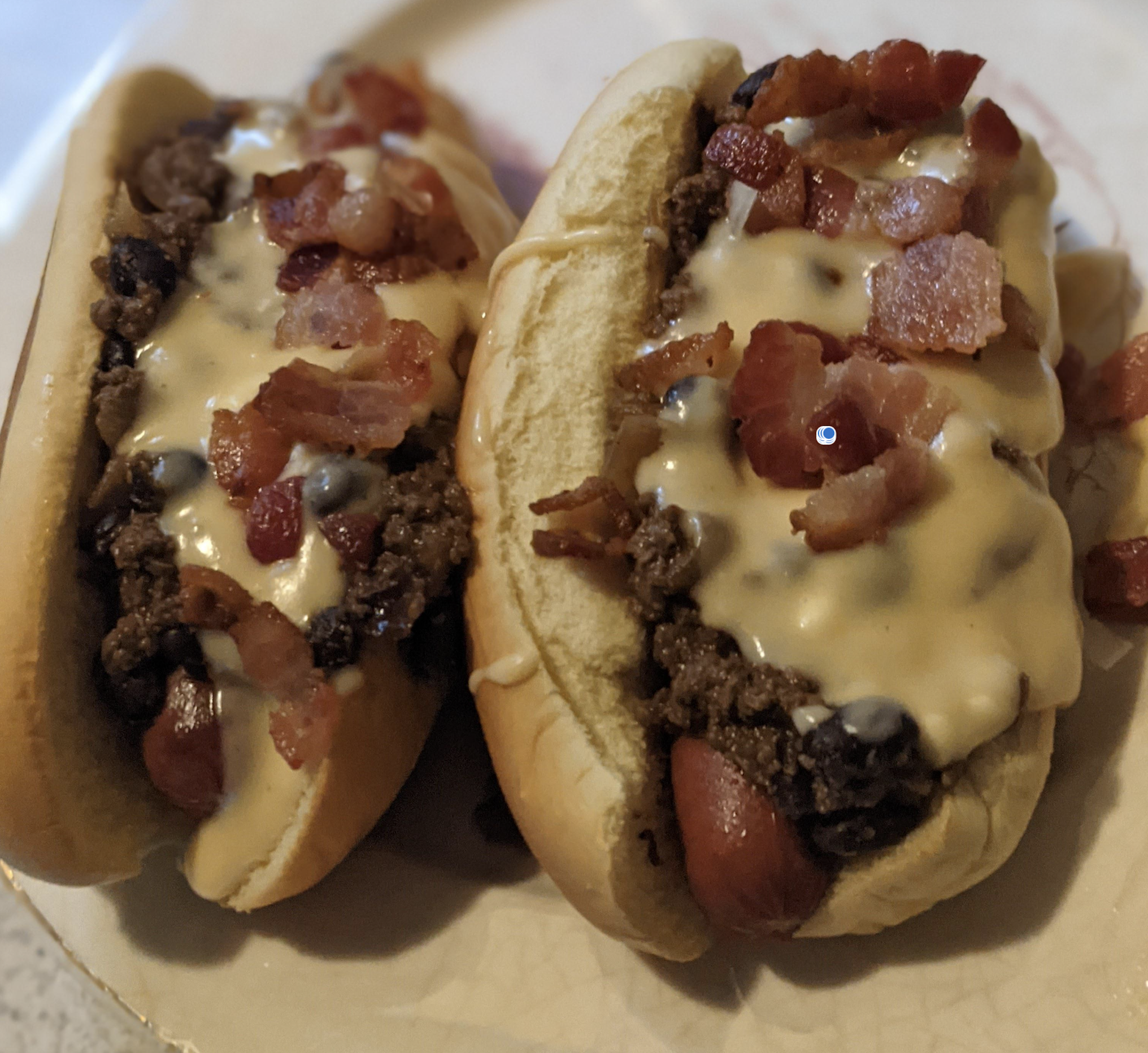 Two hot dogs in buns topped with ground beef, melted cheese, and bacon pieces, served on a light-colored plate.