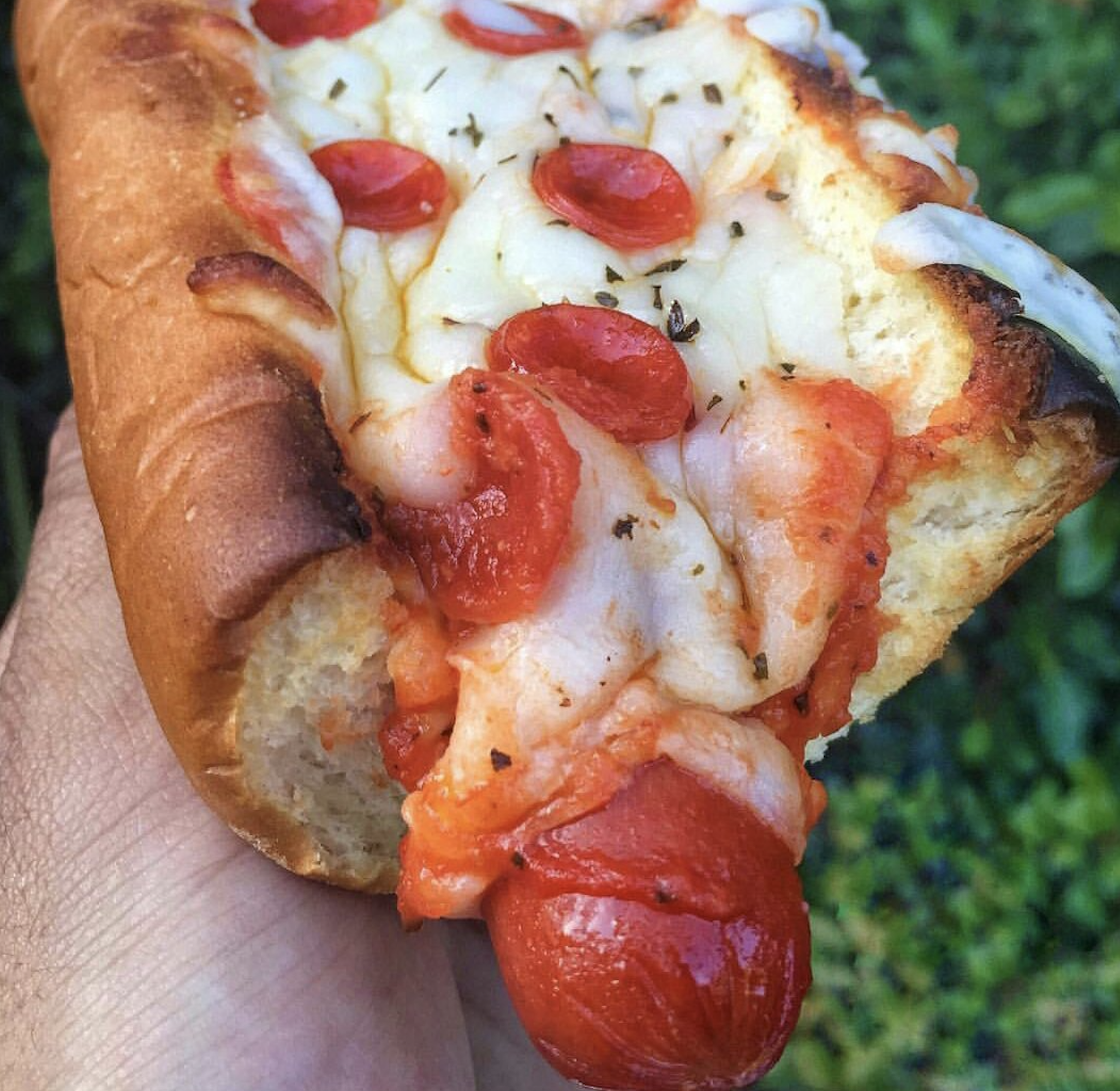 A hand holds a pizza-style hot dog. A hot dog bun is topped with tomato sauce, melted cheese, and mini pepperoni slices. The hot dog rests inside, covered in pizza toppings, set against a blurred green background.