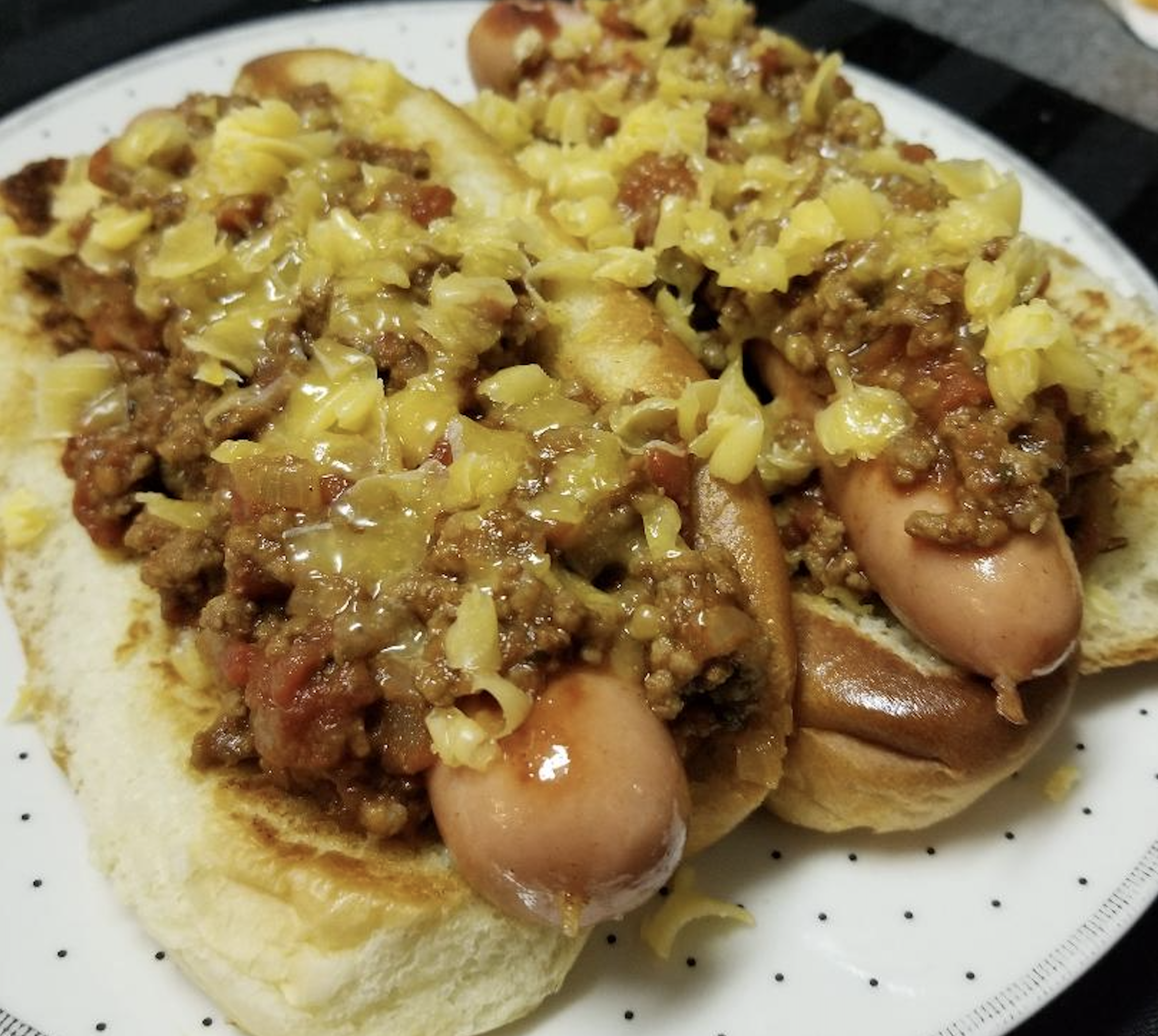 Two hot dogs in toasted buns topped with chili and shredded cheese, served on a white plate with black dots.
